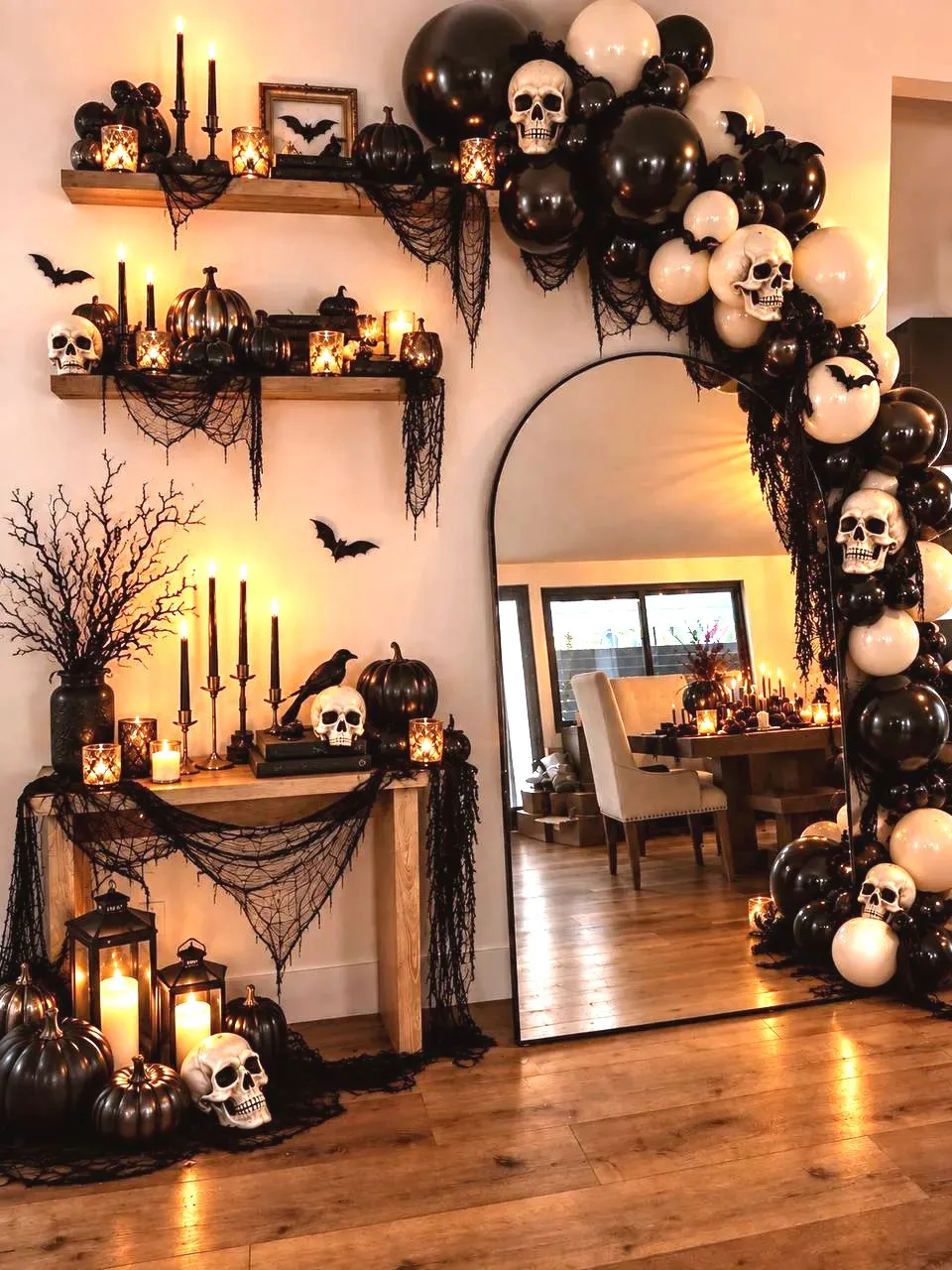 A black and cream balloon garland with skulls and bats climbing a tall arched mirror, beside wood shelves of black pumpkins, candles and draped black cheesecloth