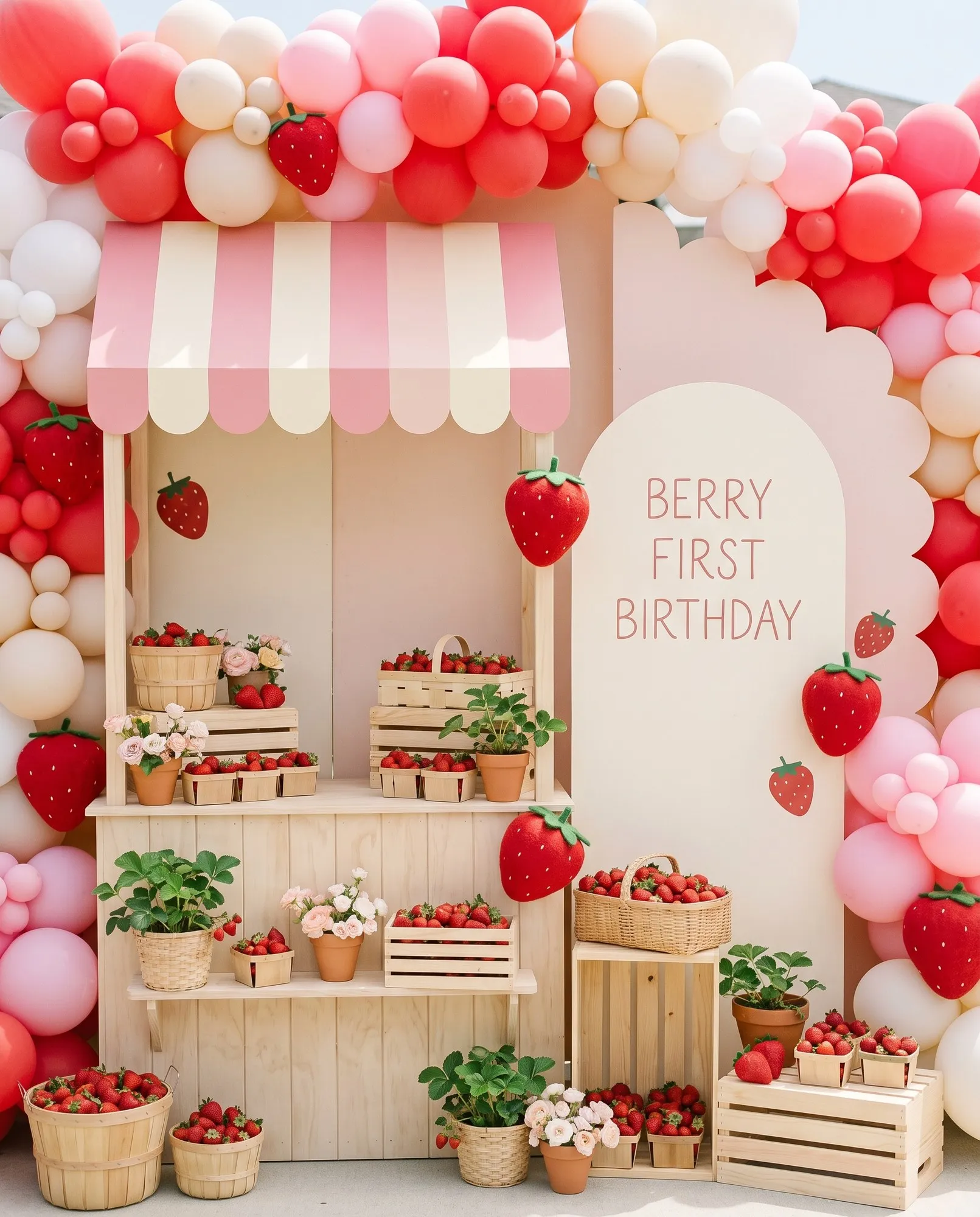 A multi-piece party backdrop — a pale wooden market stall under a scalloped pink and cream striped awning, its counter and crates crowded with baskets of fresh strawberries, potted strawberry plants and pastel blooms, standing shoulder to shoulder with a tall arched cream board hand-lettered Berry First Birthday, with blank scalloped panels overlapping at their join so the silhouette steps up and down, all of it packed around with a dense balloon installation in strawberry red, bubblegum pink, blush and cream and hung with oversized felt strawberries, the only decorative motif on the boards.
