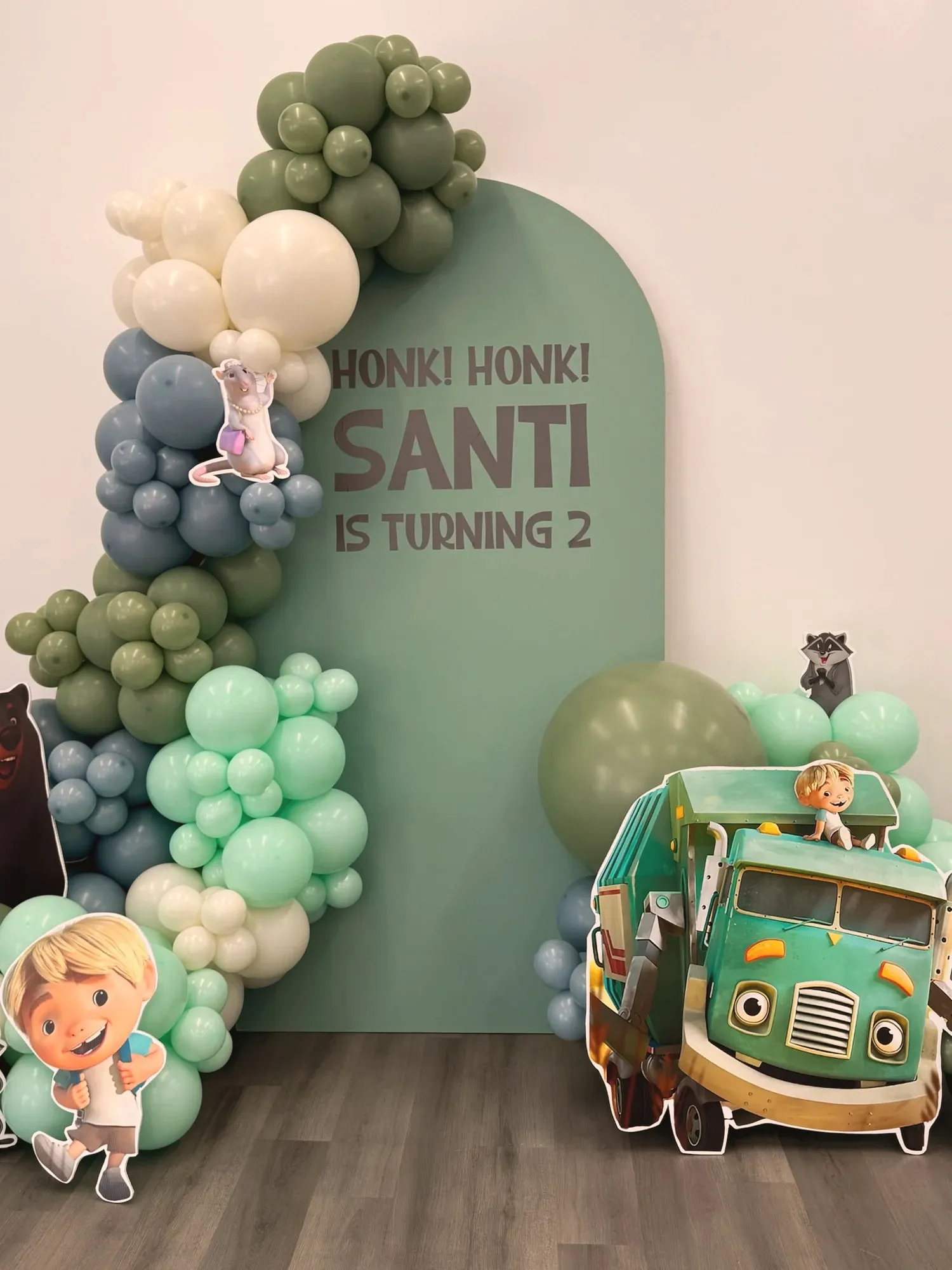 The sage arch and balloon garland with trash-truck show characters tucked into the balloons