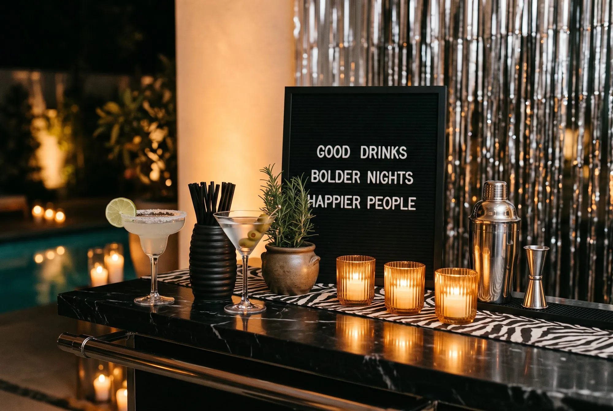 A black marble bar top by the pool with a margarita and an olive martini, a black letterboard sign reading GOOD DRINKS, BOLDER NIGHTS, HAPPIER PEOPLE, amber ribbed votives on a zebra print runner, a chrome shaker and jigger, and a silver fringe curtain behind.