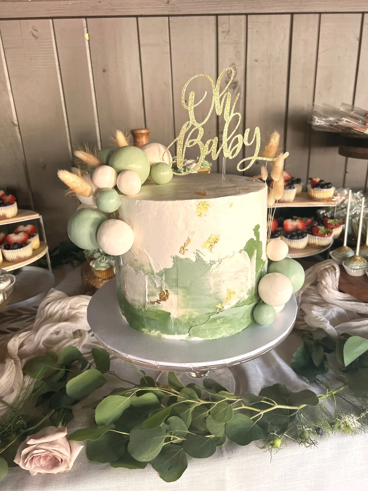 A green watercolor Oh Baby cake with gold leaf, fondant spheres, and dried pampas on a cake stand, with the dessert table behind it