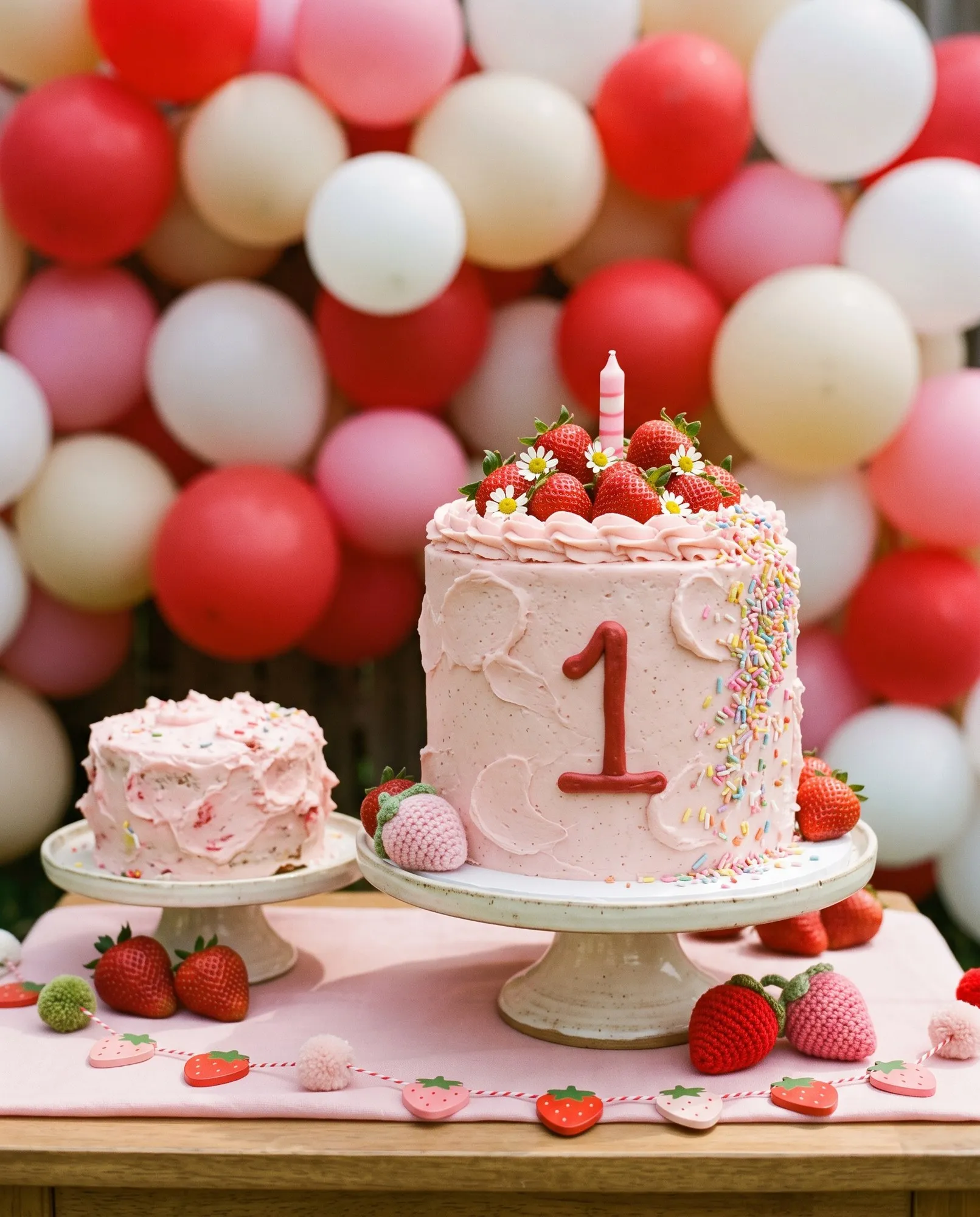 A short chunky single-tier cake in soft pink buttercream with big playful swoops and a large numeral 1 piped on the front in deep strawberry red, a thick fall of pastel rainbow sprinkles down one side, whole strawberries and small white daisies piled on top and one stubby striped candle, beside a deliberately messy little plain smash cake, both in front of a dense wall of red, pink and cream balloons.