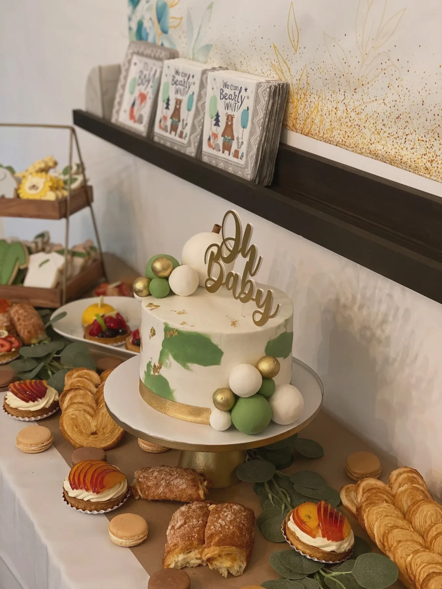 A close-up of the sage and gold Oh Baby cake topped with fondant spheres, surrounded by macarons and pastries