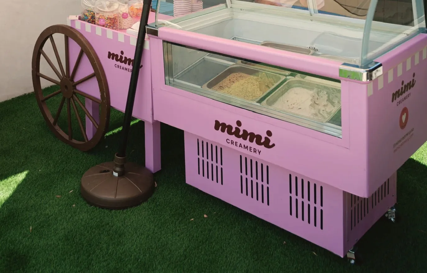 A pale pink ice cream cart with a wooden spoked wheel and a curved glass display case, holding two tubs of gelato, parked on the lawn under a cream umbrella