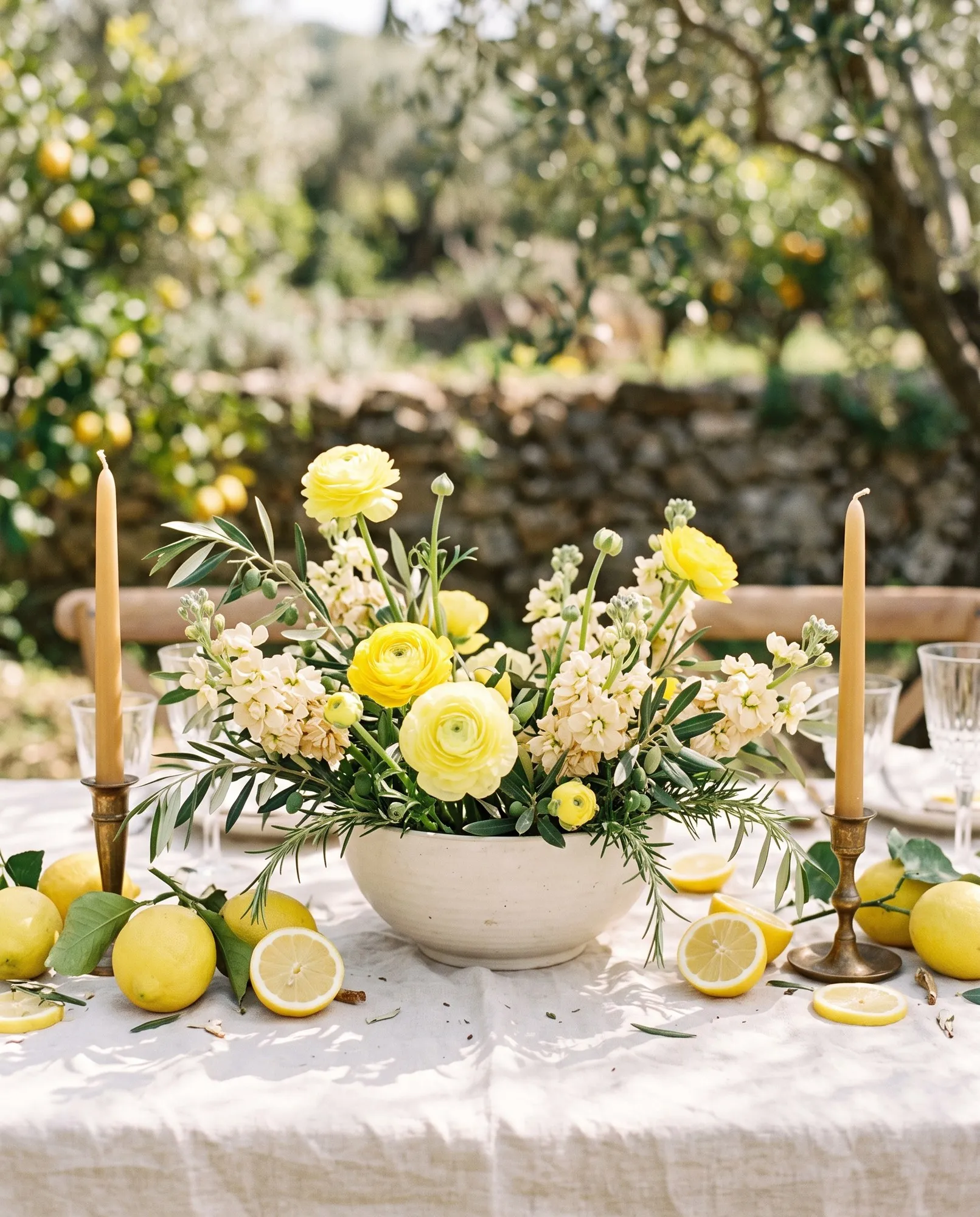 A loose arrangement of yellow ranunculus, cream stock, olive branches and rosemary in a wide unglazed cream bowl, ringed with whole and halved lemons between two honey-toned taper candles in bronze holders.