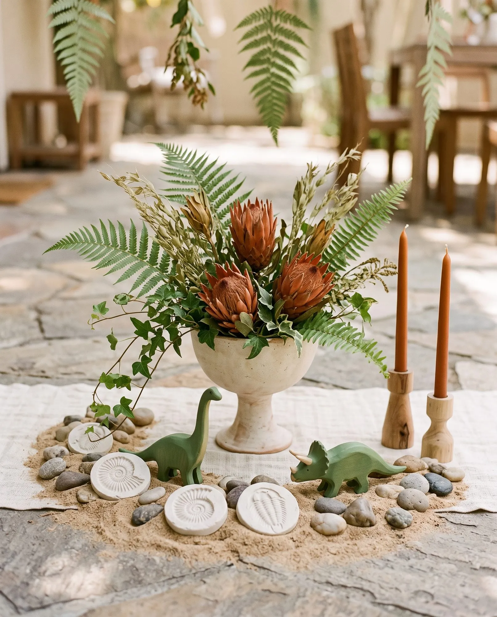 A footed cream stoneware bowl on a linen runner holding three rust proteas with dried grasses, seed heads, real fern fronds and trailing ivy spilling over one side, ringed at its base by a circle of real sand studded with smooth river stones, four white unglazed clay discs pressed with ammonite and trilobite fossils, and two green wooden dinosaurs walking through it, with a pair of rust taper candles in turned wood holders to one side and hanging paper fern fronds and blurred wooden patio furniture behind.