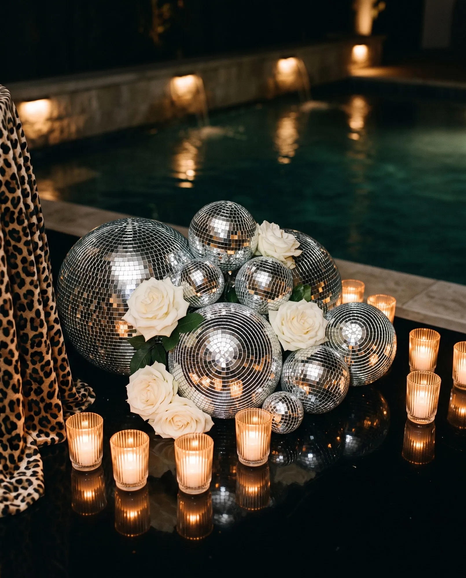 A cluster of mirror balls in six or seven different sizes grouped on a black surface with white roses tucked between them, ribbed amber votives in a line across the front and a leopard print drape falling in at the edge of frame.
