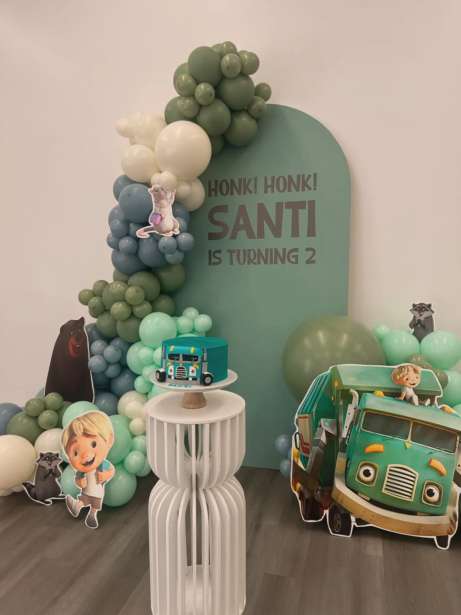 The turquoise truck cake on a white pedestal beside the matching truck cutout, in front of the sage Honk Honk Santi is Turning 2 arch and balloon garland