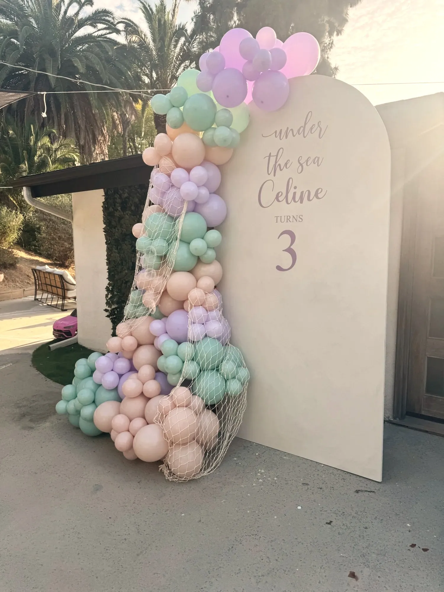 A cream arched sign reading Under The Sea, Celine Turns 3, banked with a lavender, mint and peach balloon garland draped in fishing net, with palm trees and low afternoon sun behind it