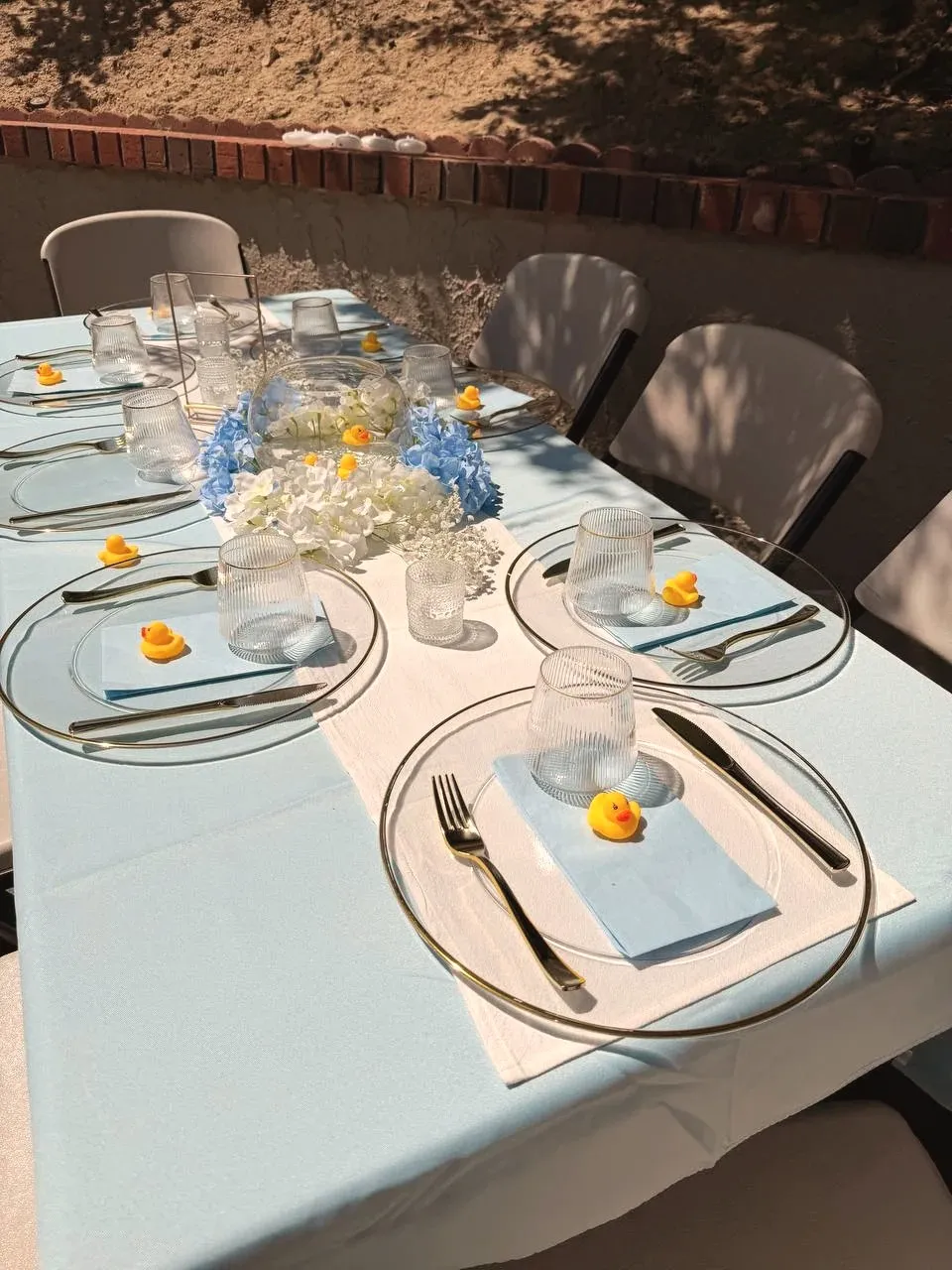 A long table dressed in pale blue, set with glass chargers, blue napkins and a small yellow duck at every place.