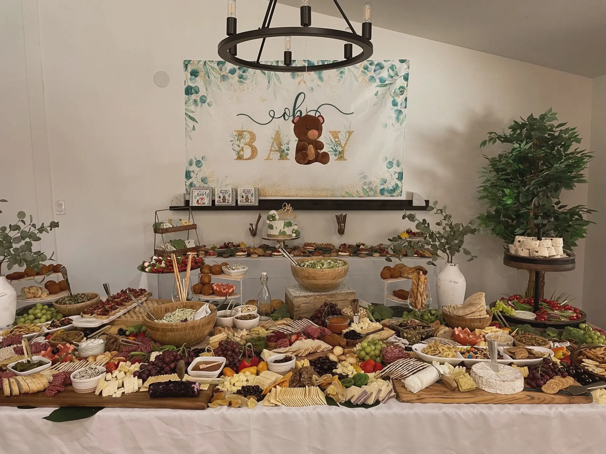 A long charcuterie grazing table piled with meats, cheeses, fruit, and breads under an Oh Baby bear backdrop and a black ring chandelier