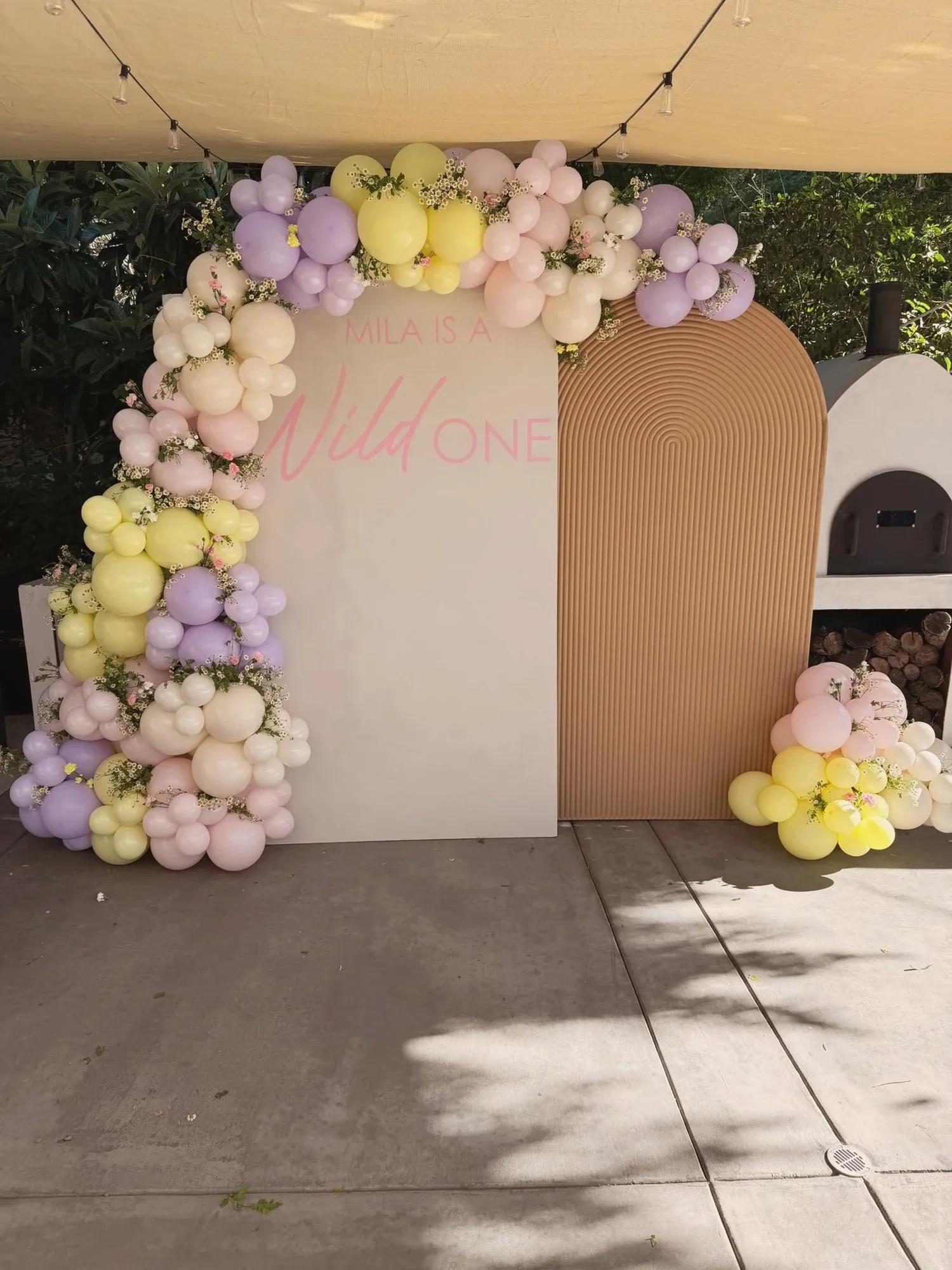 A cream arched backdrop reading Mila Is A Wild One in pink script, framed by a balloon garland in lilac, butter yellow, blush and cream with sprigs of wildflowers tucked through it