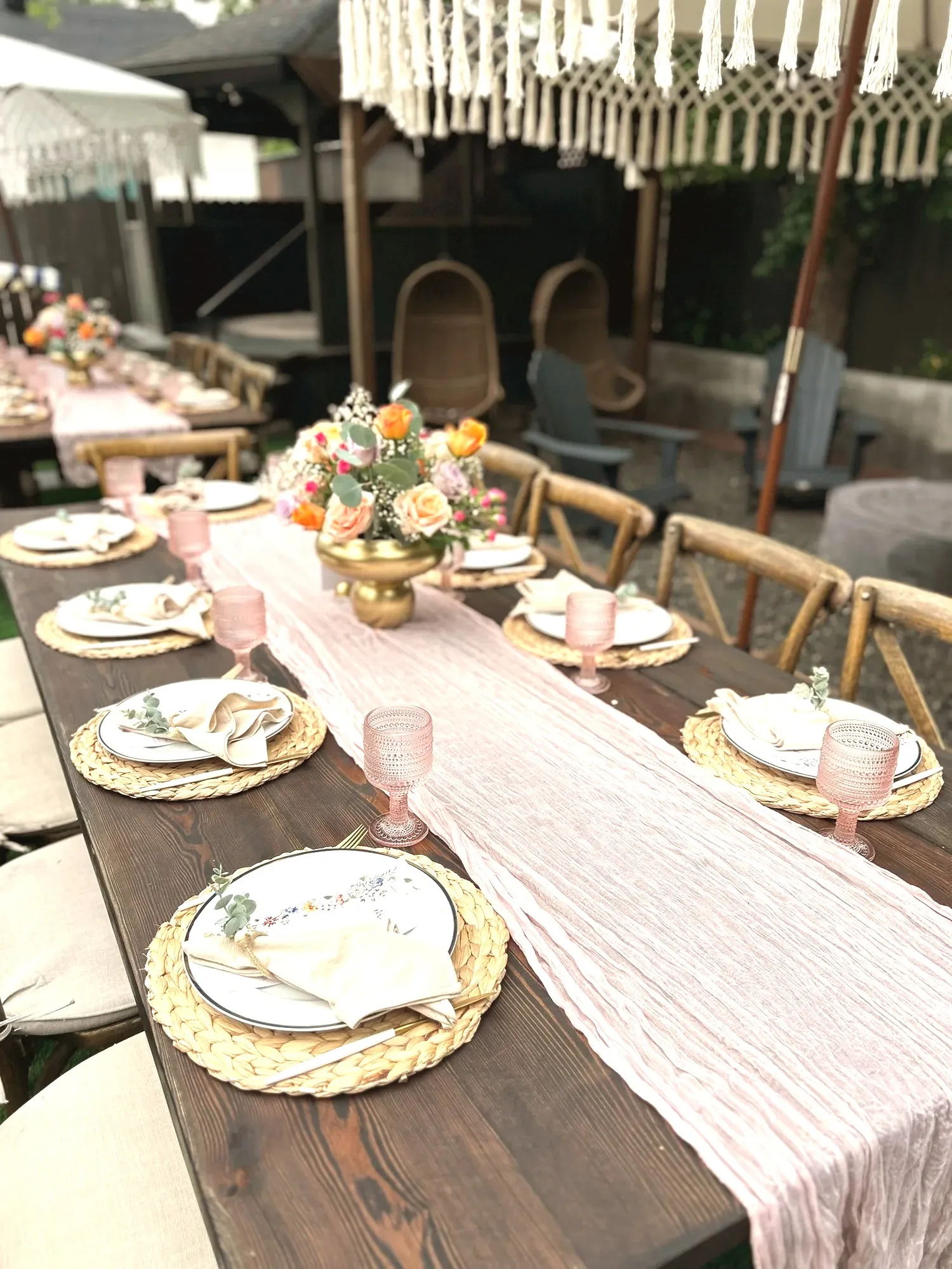 A long wood farm table with woven placemats, cream napkins and pink glassware, a dusty pink runner down the middle and a low gold bowl of orange, peach and lilac roses