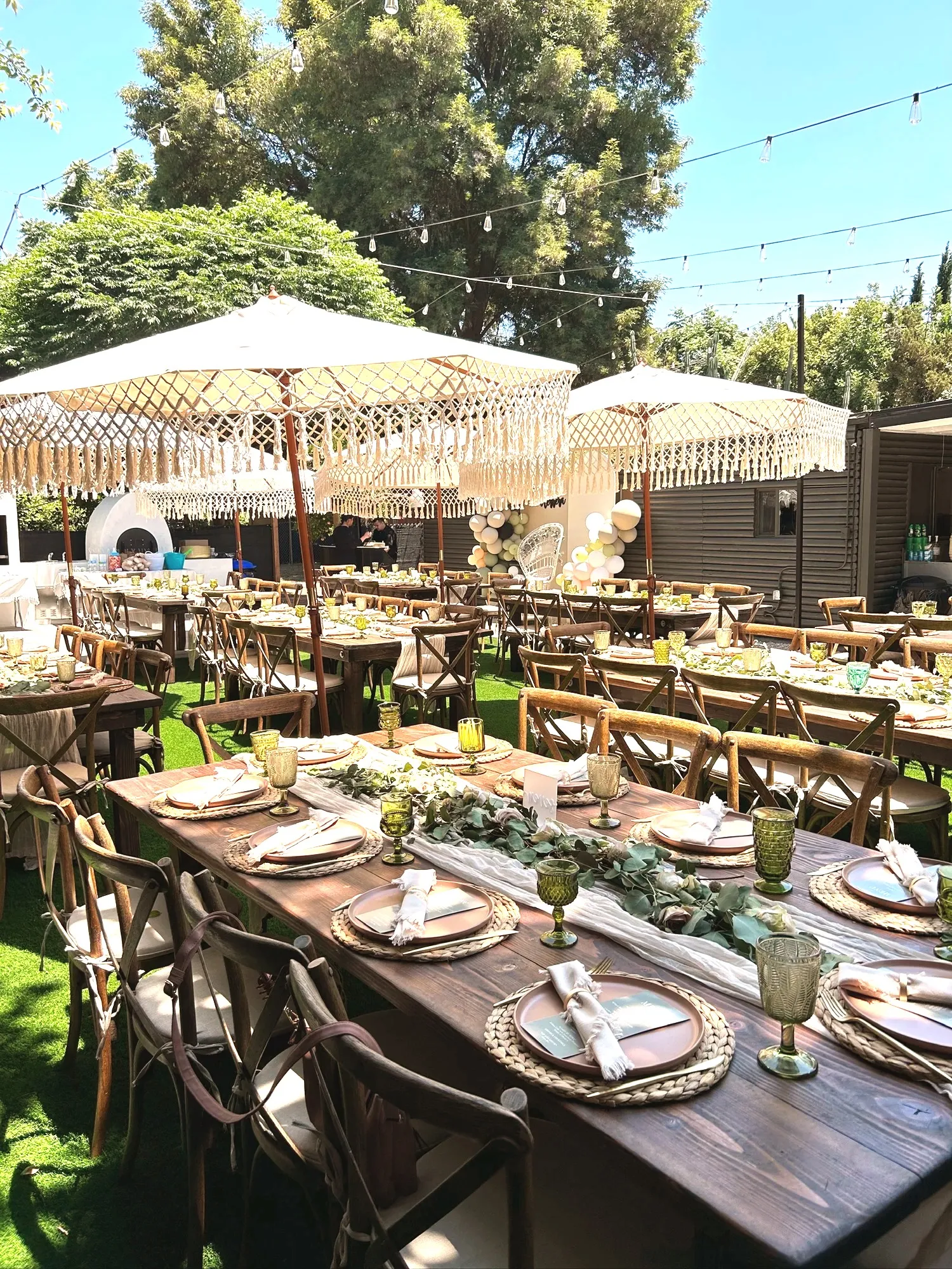 A large backyard shower seen from above, long wood farm tables set with terracotta plates and eucalyptus runners under cream macramé-fringed umbrellas, with string lights overhead