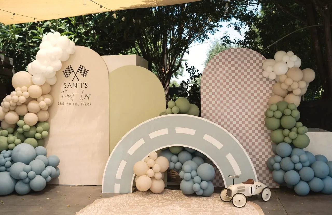 A racing birthday backdrop of layered arched boards, a checkered arch, and a curved race track, banked with cream, sage, and blue balloons