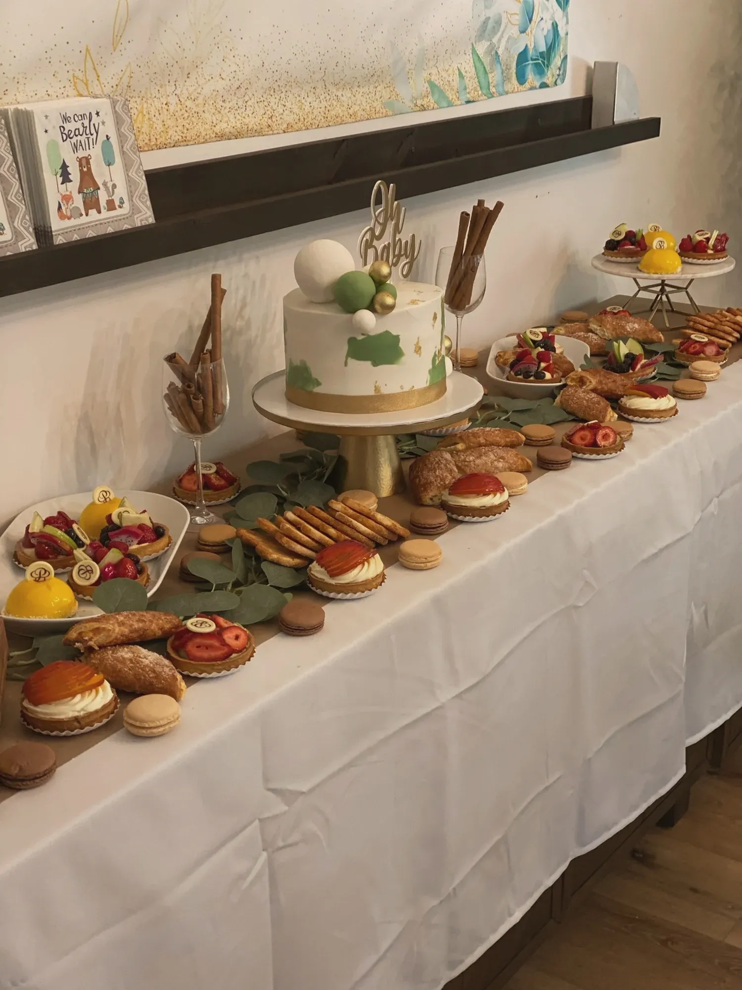 A woodland dessert table with a green and gold Oh Baby cake, fruit tarts, macarons, and we-can-bearly-wait napkins
