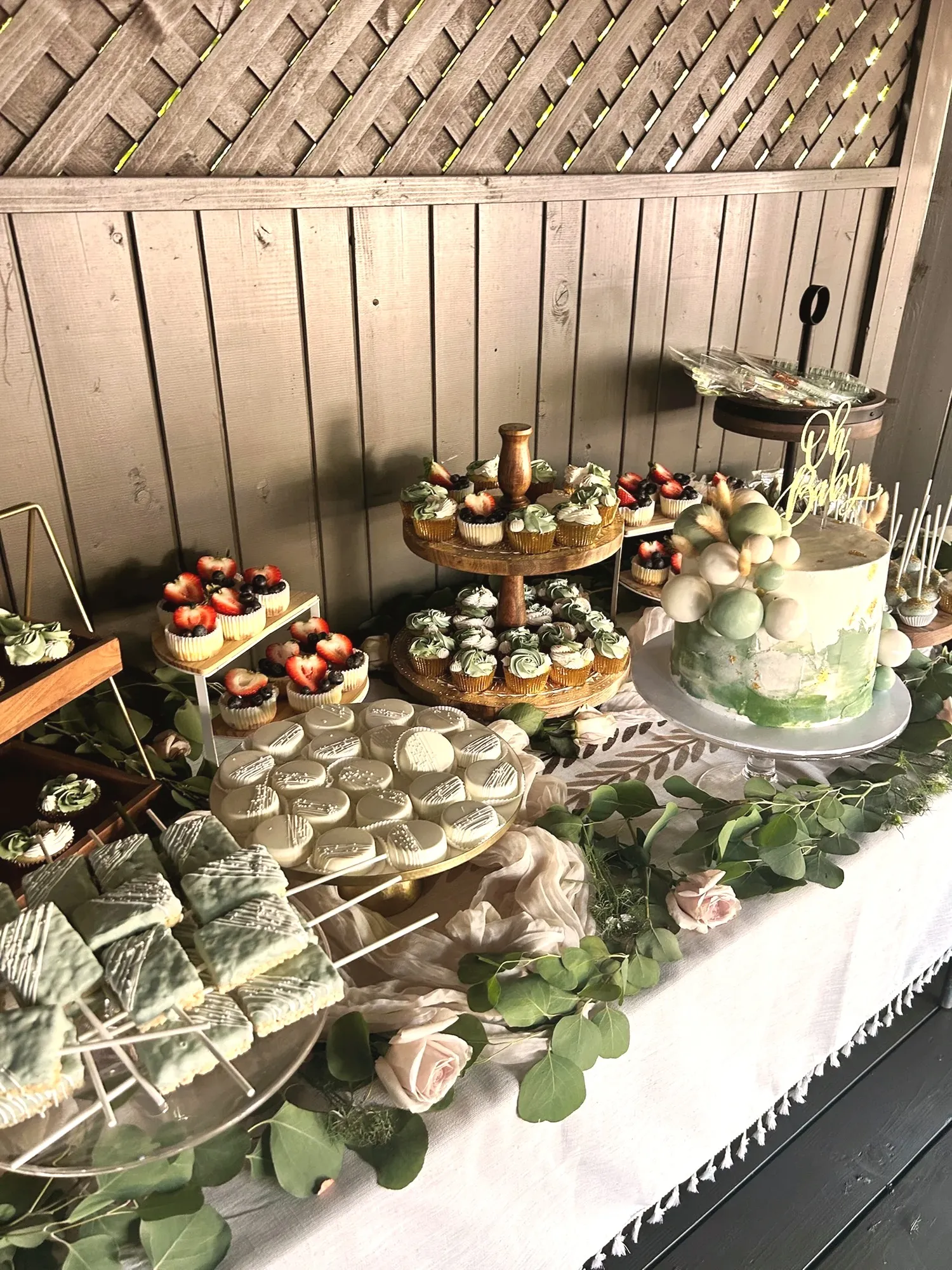 A dessert table with green-iced cupcakes, chocolate-dipped cookies, mini cheesecakes with berries, cake pops, and the green Oh Baby cake, trailed with eucalyptus and blush roses