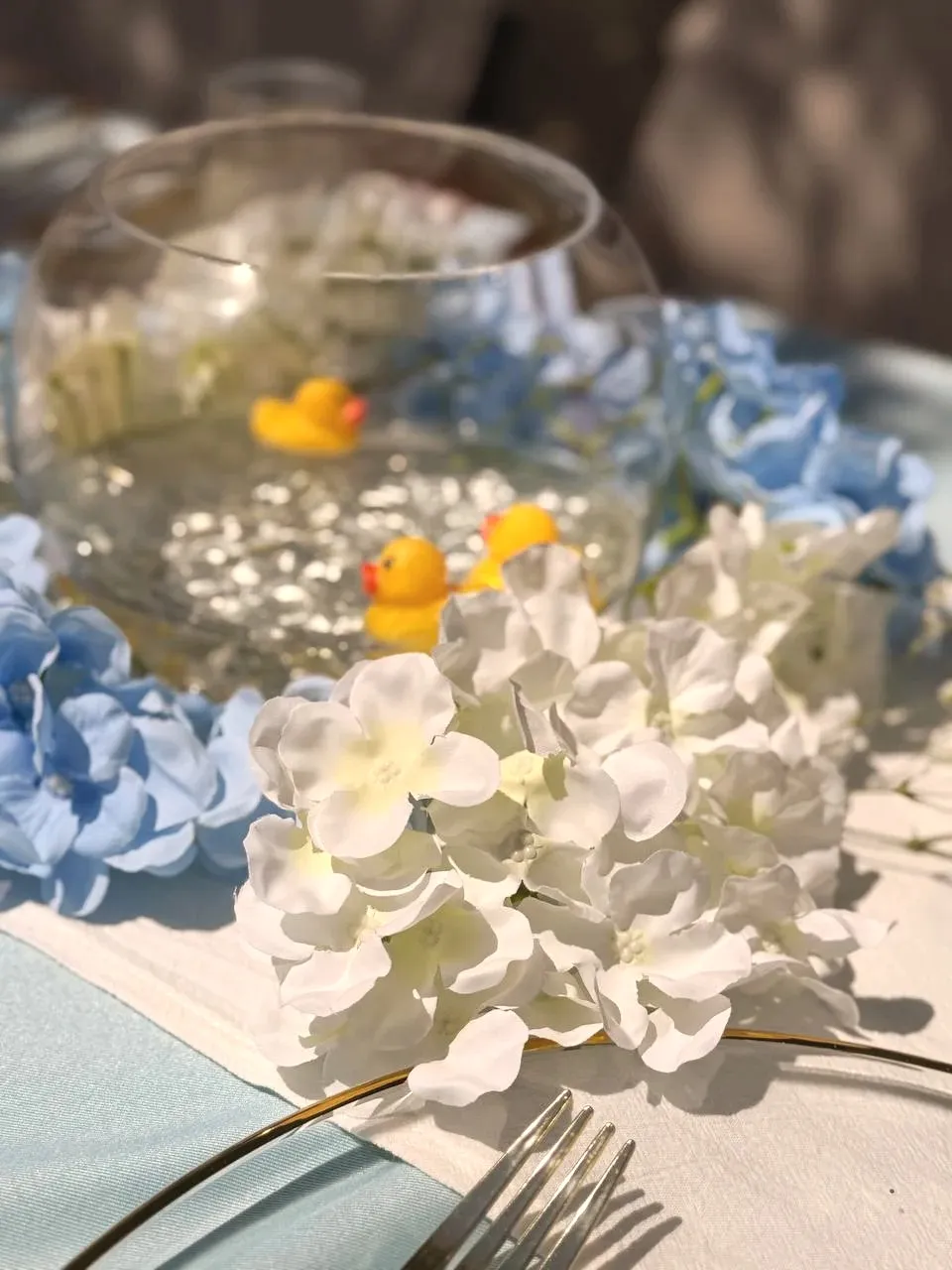 White hydrangea blooms in the foreground, with rubber ducks floating in a glass bowl behind them.