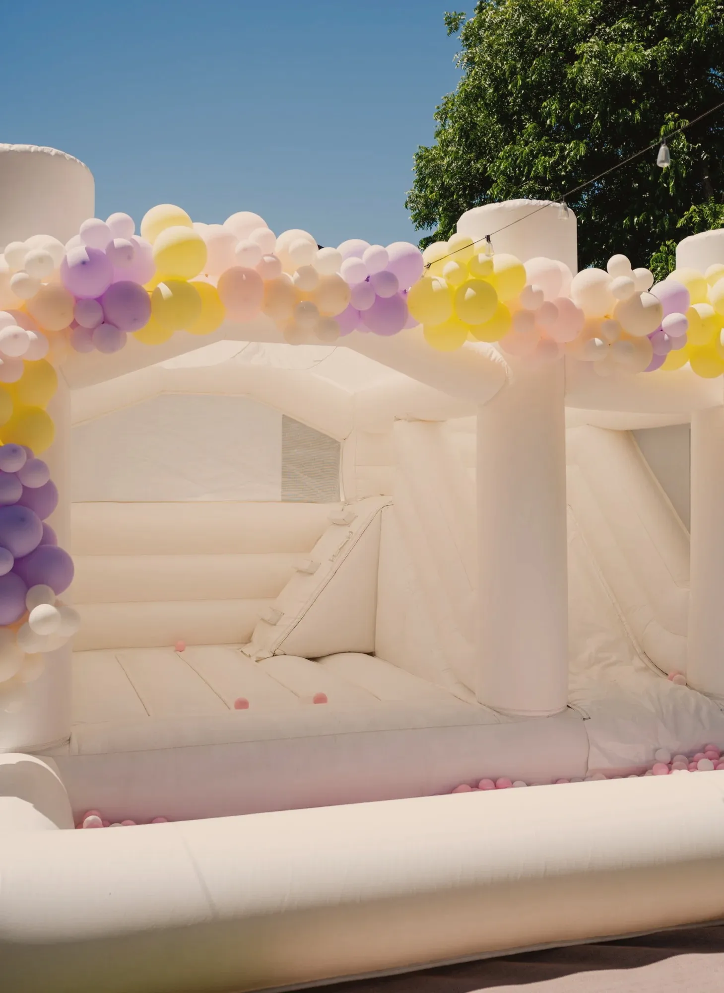 A white bounce house with a slide and a ball pit of pale pink balls, topped with a garland of lilac, butter yellow, blush and cream balloons against a clear blue sky