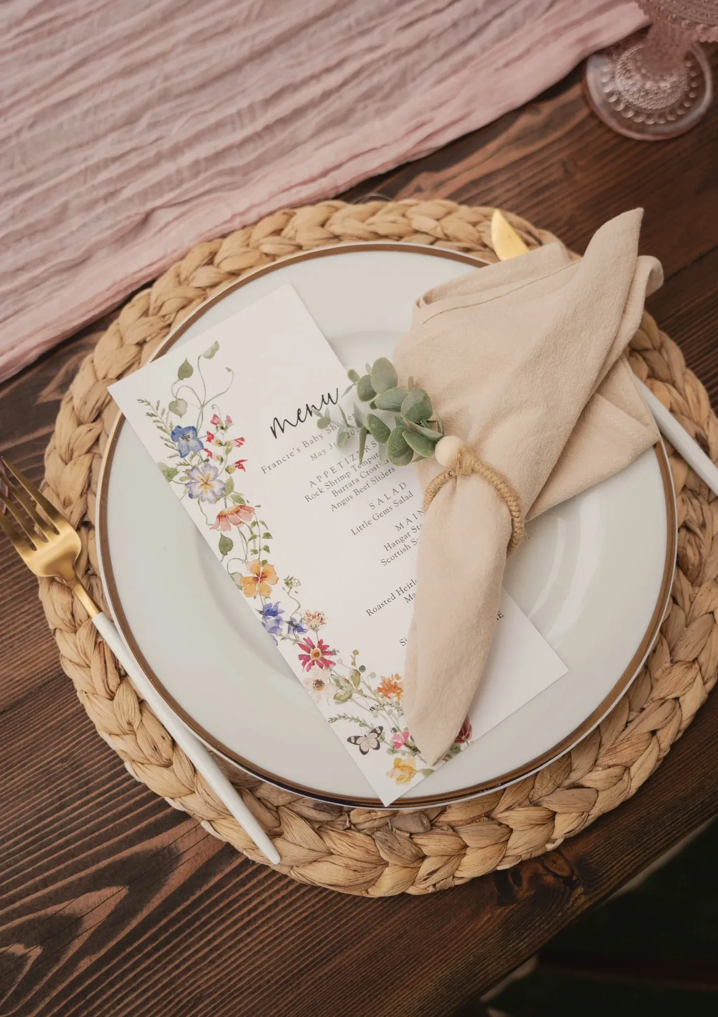 A place setting on a woven mat, gold-rimmed plate with a wildflower menu card, a cream napkin in a beaded ring with a sprig of eucalyptus, on a dusty pink runner