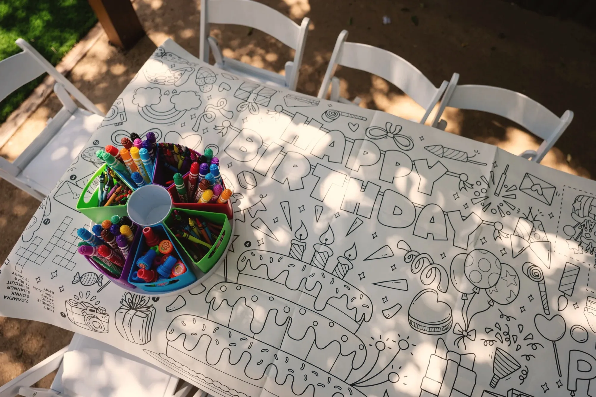 A children's table covered in a black and white Happy Birthday coloring sheet with a carousel of markers, crayons and stampers in the middle, dappled with shade