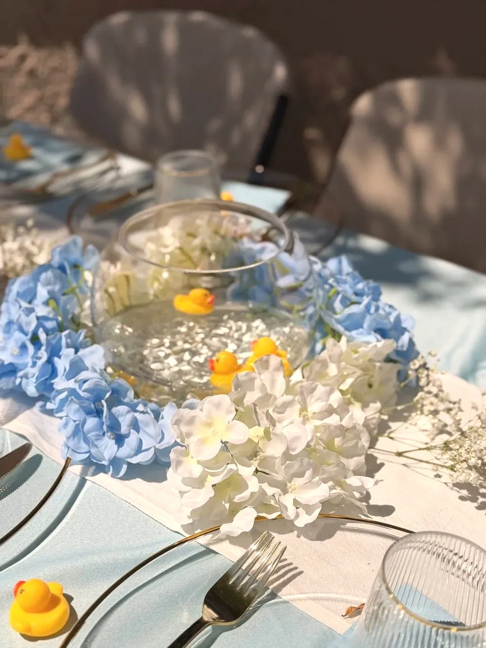 A glass fishbowl of water with three yellow ducks in it, ringed with blue and white hydrangea.