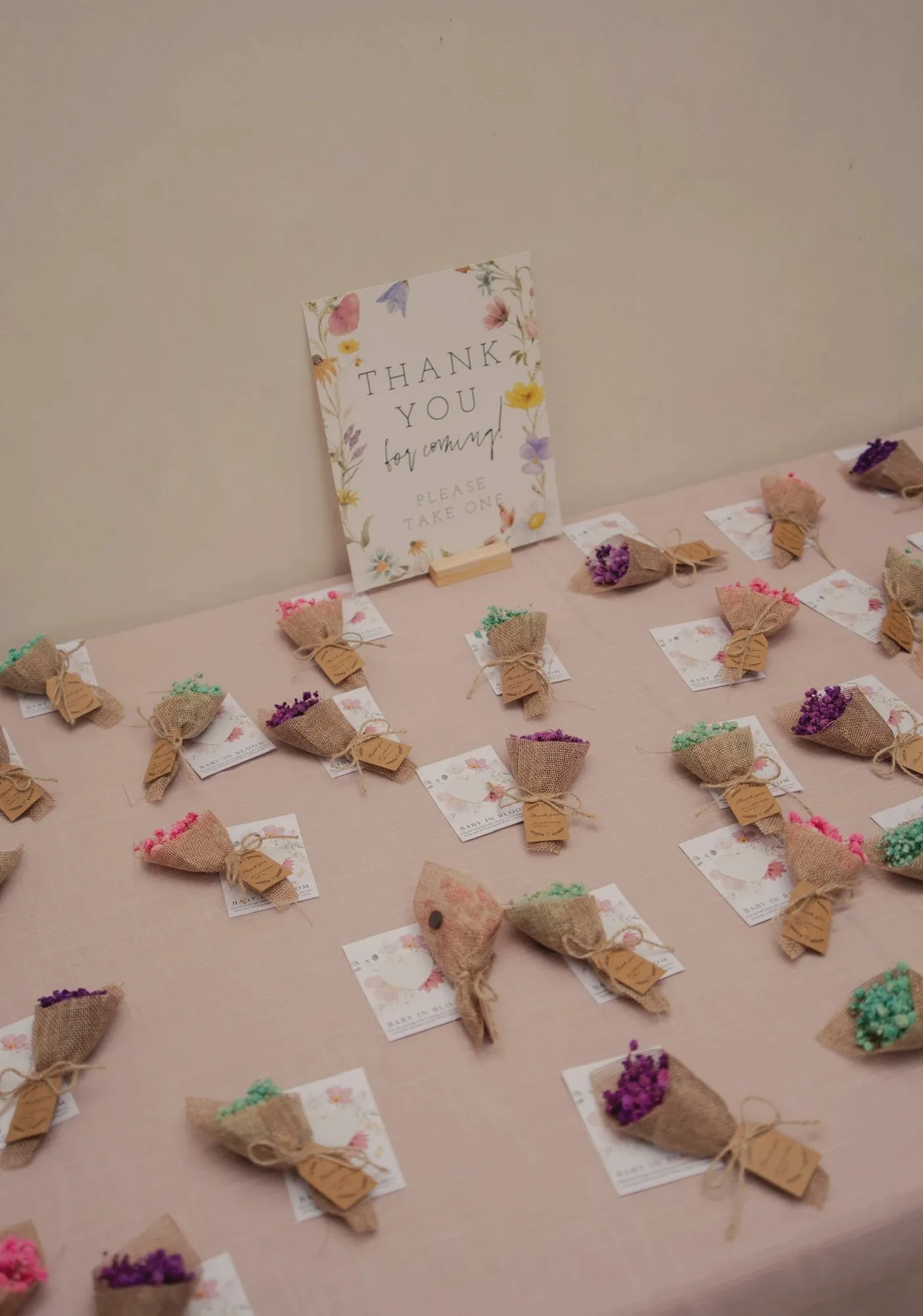 Rows of mini dried-flower bouquets wrapped in burlap cones with kraft tags, laid out on a pink cloth beside a floral thank-you-for-coming sign