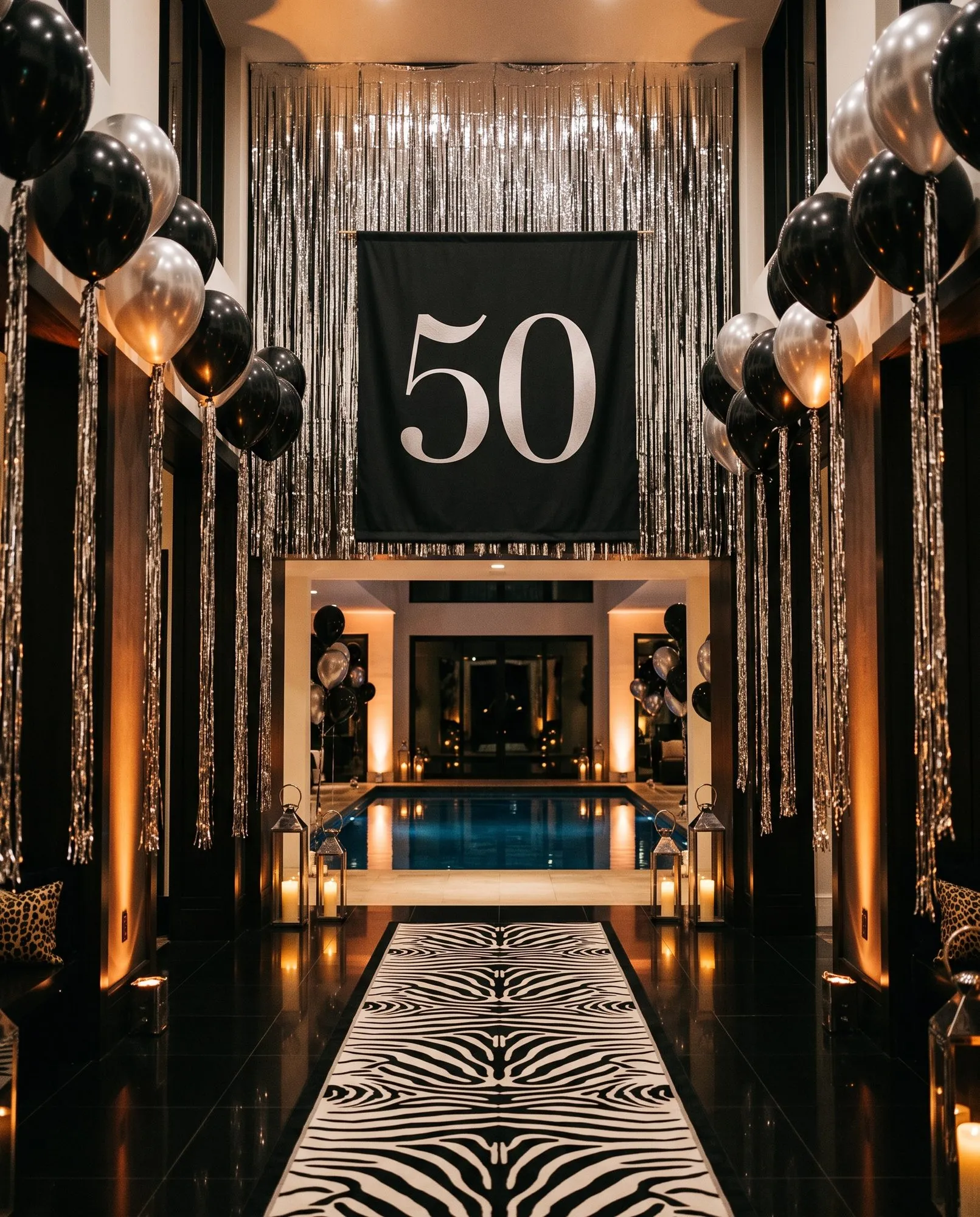 A hallway lined with columns of black and chrome balloons trailing silver fringe, a zebra print runner down the dark tile floor, and a black banner printed with a silver 50 hung against a wall of metallic fringe, with a candlelit pool visible through the far doorway.