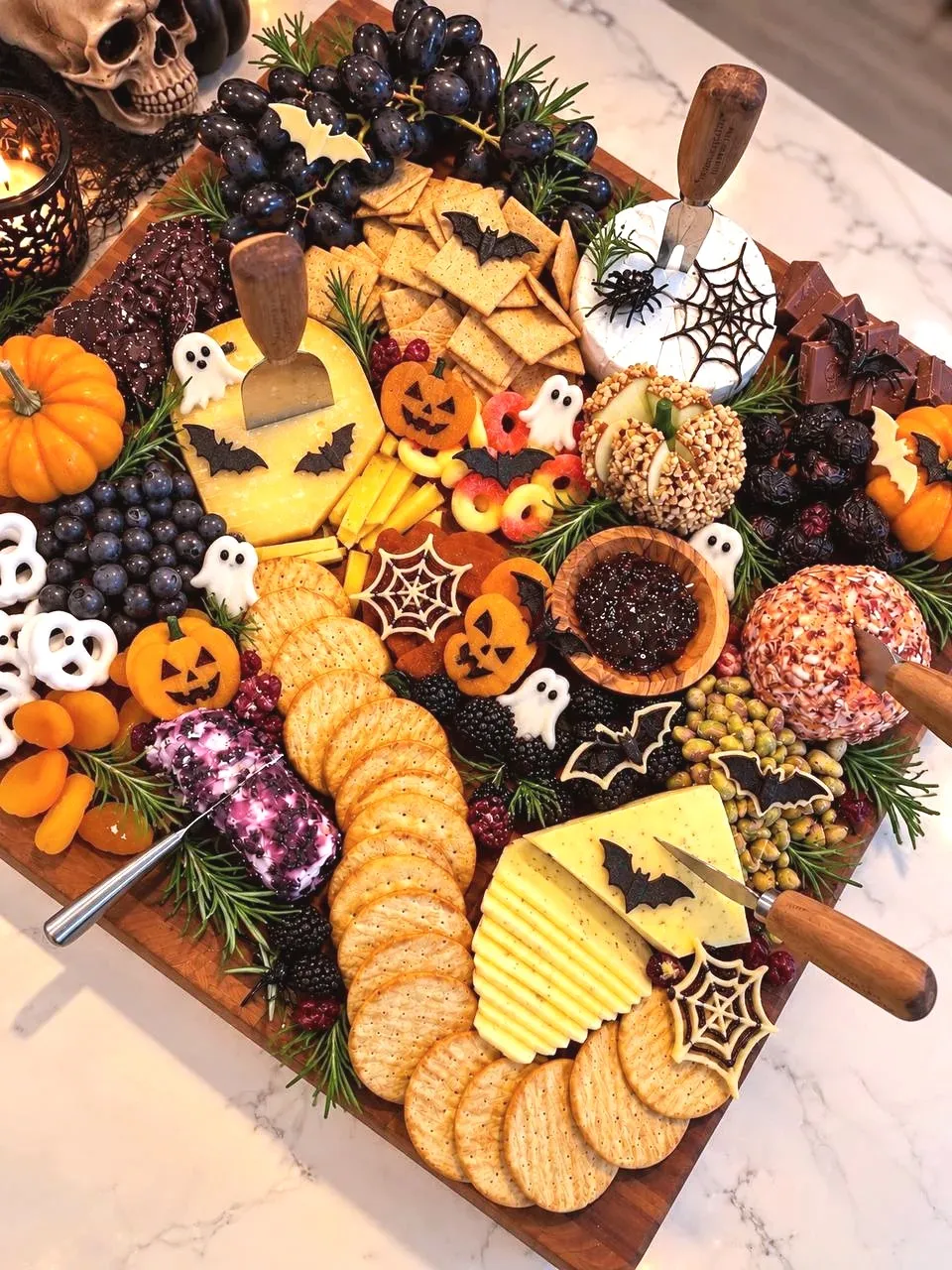A Halloween grazing board with black grapes, crackers, brie under a chocolate spiderweb, cheese cut with bats, mini pumpkins, white chocolate ghosts and gummy rings