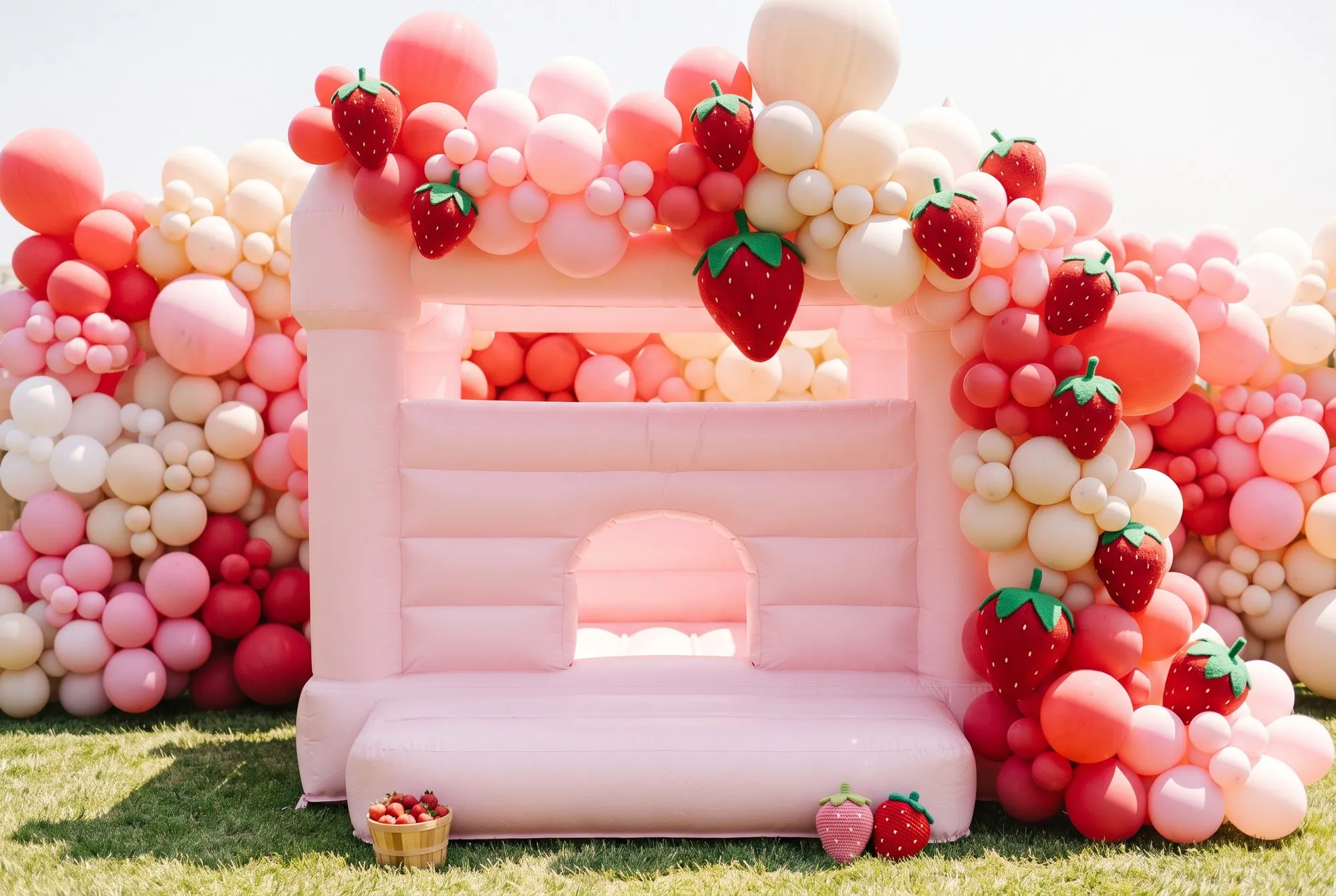 A plain light pink inflatable bounce house with flat blush panels, a square top and a low arched entrance, undecorated except for a lavish organic balloon garland in the same strawberry red, bubblegum pink, blush and cream as the backdrop, sweeping across the top and cascading down one side with oversized felt strawberries worked into it, and a woven basket of strawberries and two crocheted toy strawberries set at the entrance.