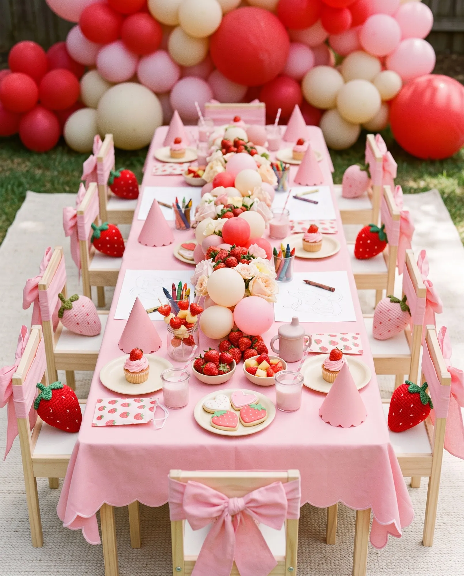 A long toddler-height table under a candy-pink scalloped cloth, tiny pale wooden chairs each tied with a fat pink bow, a low centerpiece of strawberries, mini balloons and pastel blooms running the length of it, and every place set for a small child with a pink paper party hat, a frosted mini cupcake, shaped sugar cookies, bowls of strawberries, cups of strawberry milk with striped straws, crocheted toy strawberries and pots of chunky crayons.