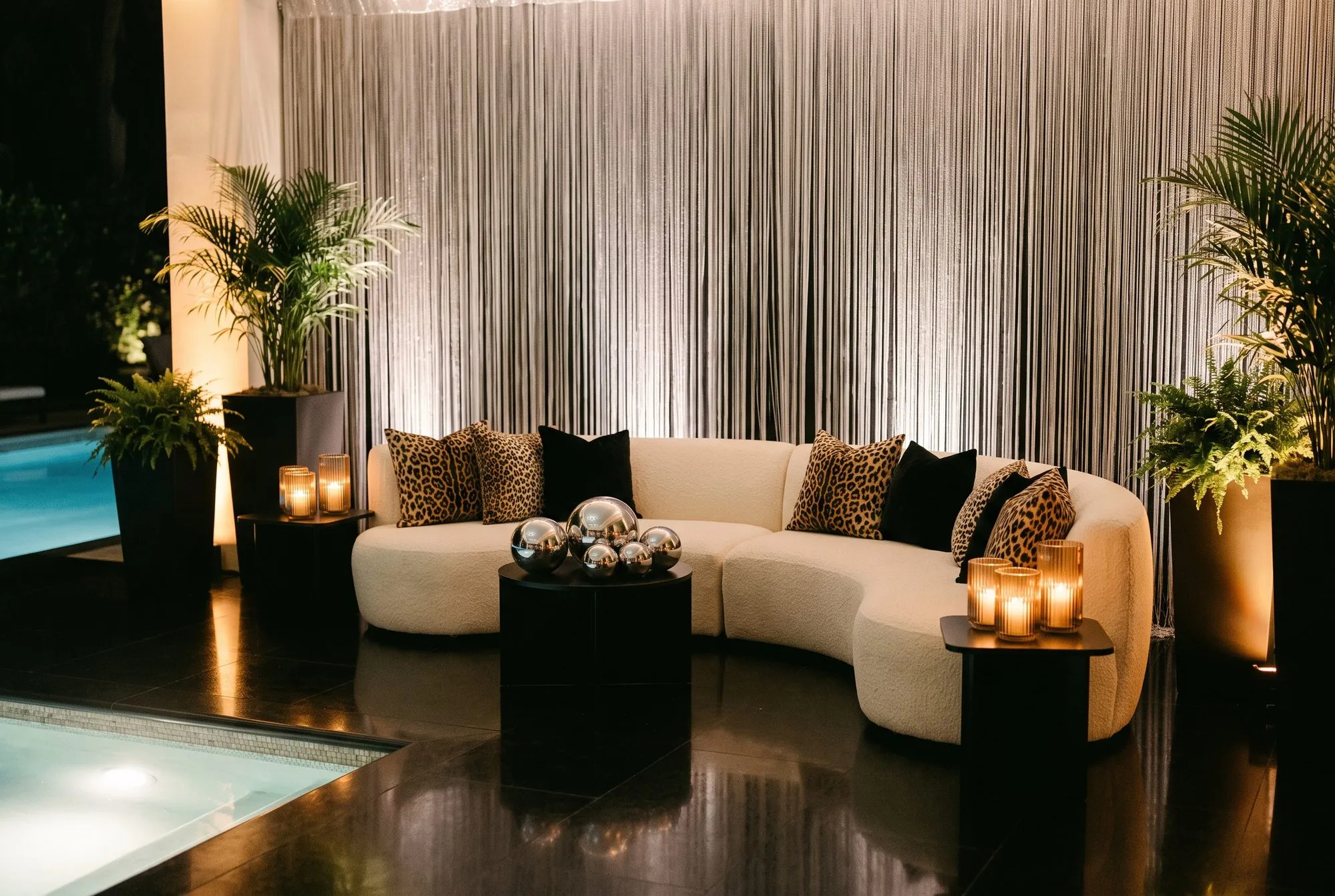 A curved cream boucle sofa against a floor to ceiling silver fringe wall, piled with leopard print and black velvet cushions, a black round table holding a cluster of chrome spheres, potted palms and ferns, and candles in ribbed glass on either side.