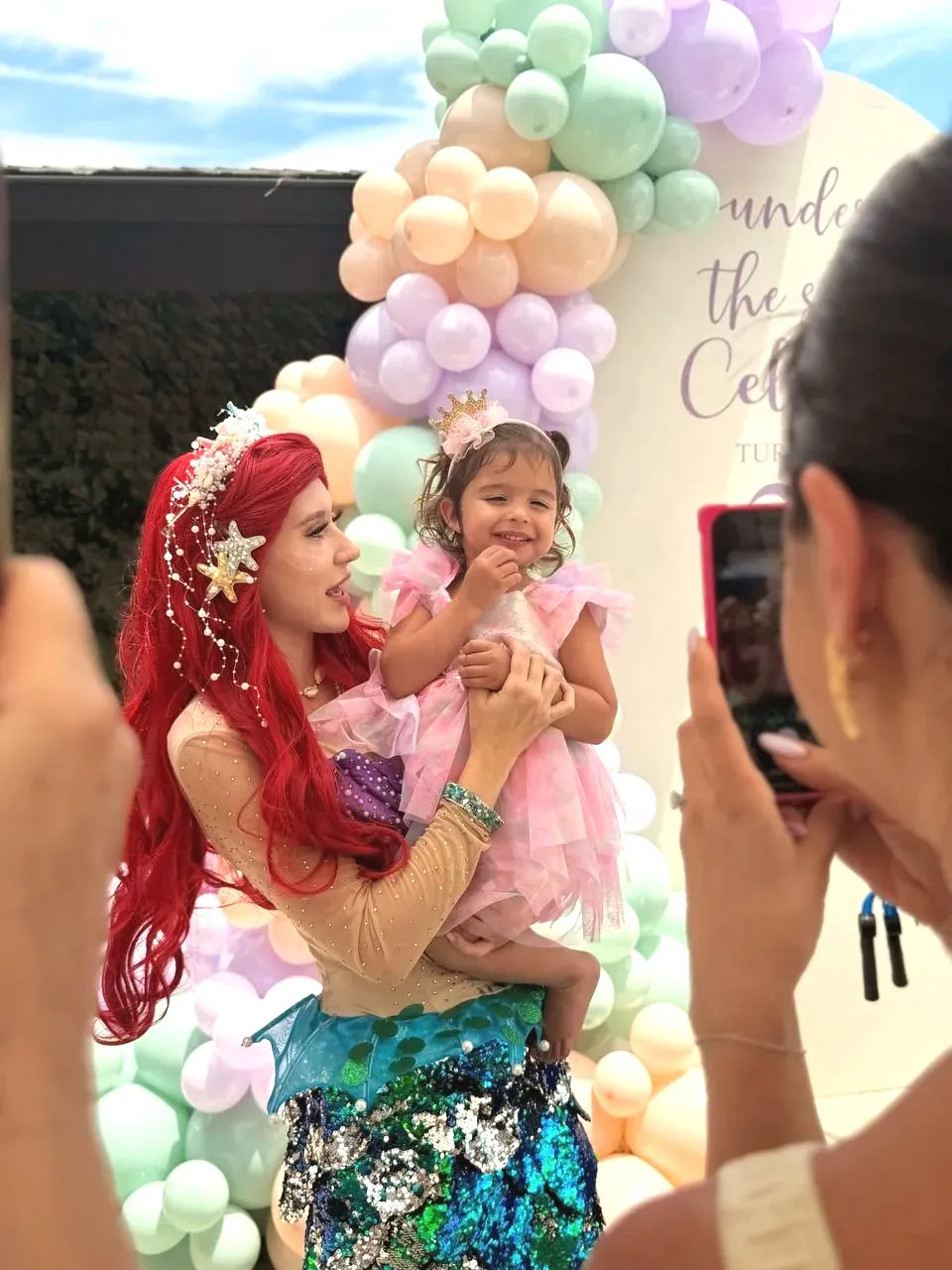 The mermaid holding the birthday girl in a pink tulle dress and a shell crown in front of the balloon arch, while a guest photographs them