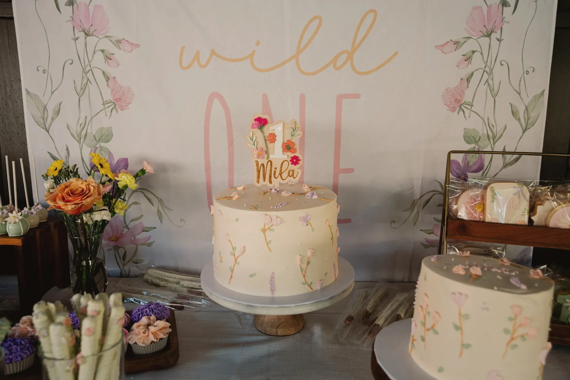 A cream buttercream cake hand-painted with wildflower stems, topped with a wooden number one and Mila's name, in front of a painted floral Wild One banner with cupcakes and cookies beside it