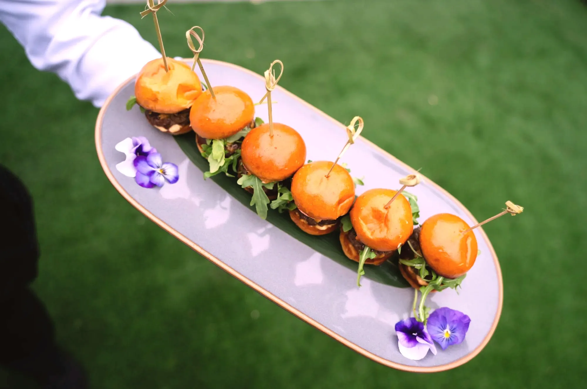 A tray of angus beef sliders on brioche buns with arugula and bamboo picks, garnished with purple pansies