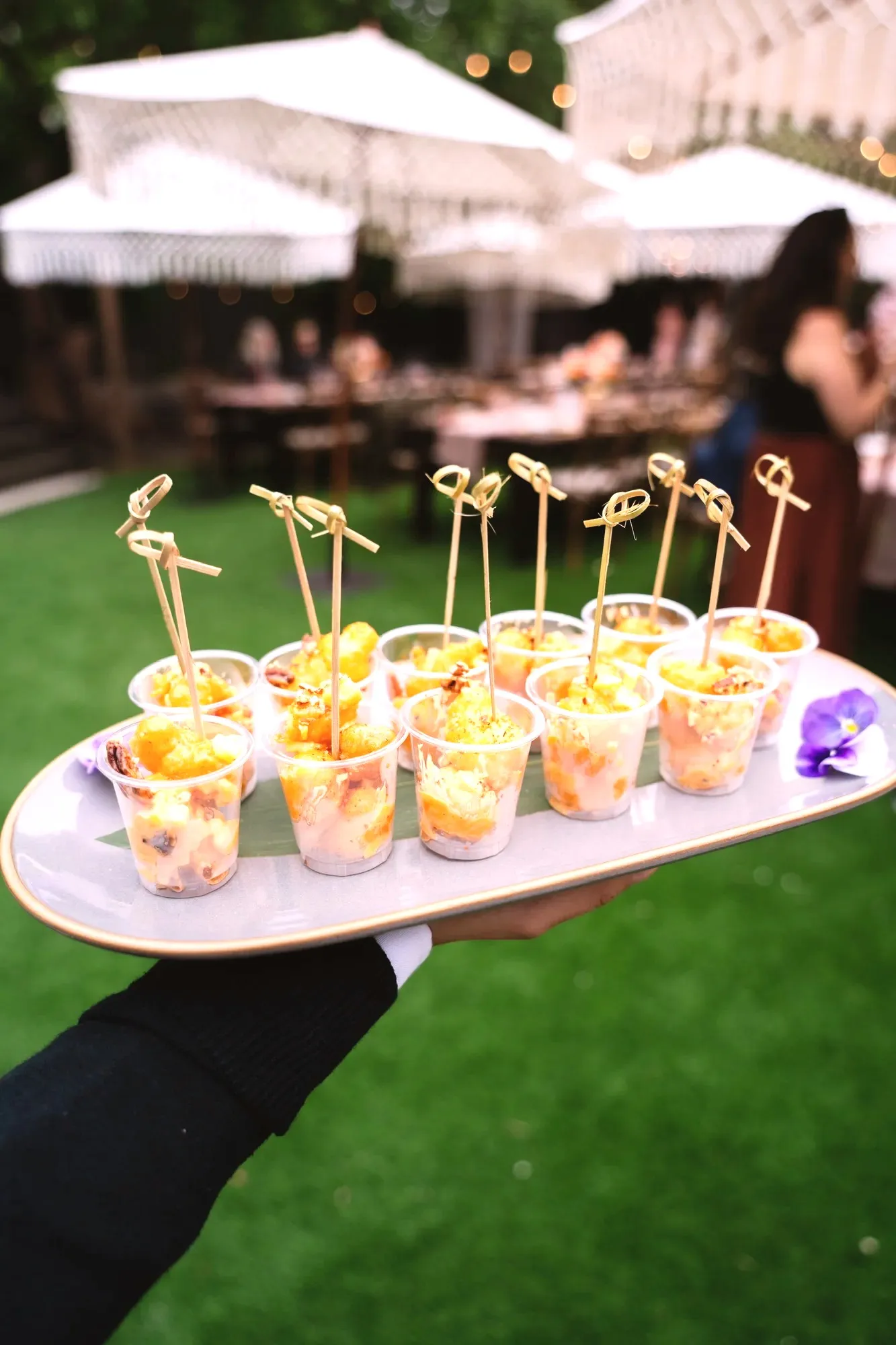 A tray of rock shrimp tempura cups with bamboo picks, held over the lawn with the macramé umbrellas behind