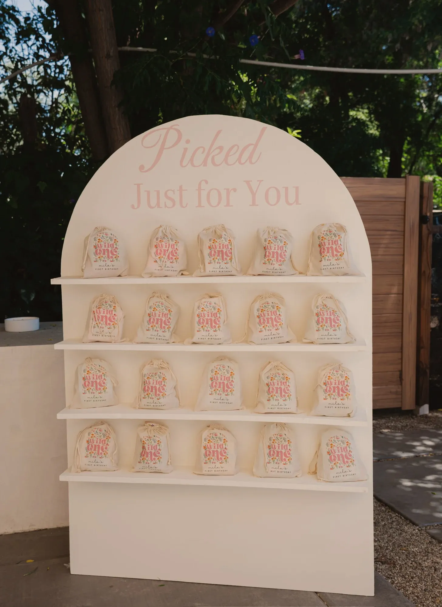 A cream arched favour stand reading Picked Just For You, holding twenty canvas drawstring bags printed with a wildflower Wild One design