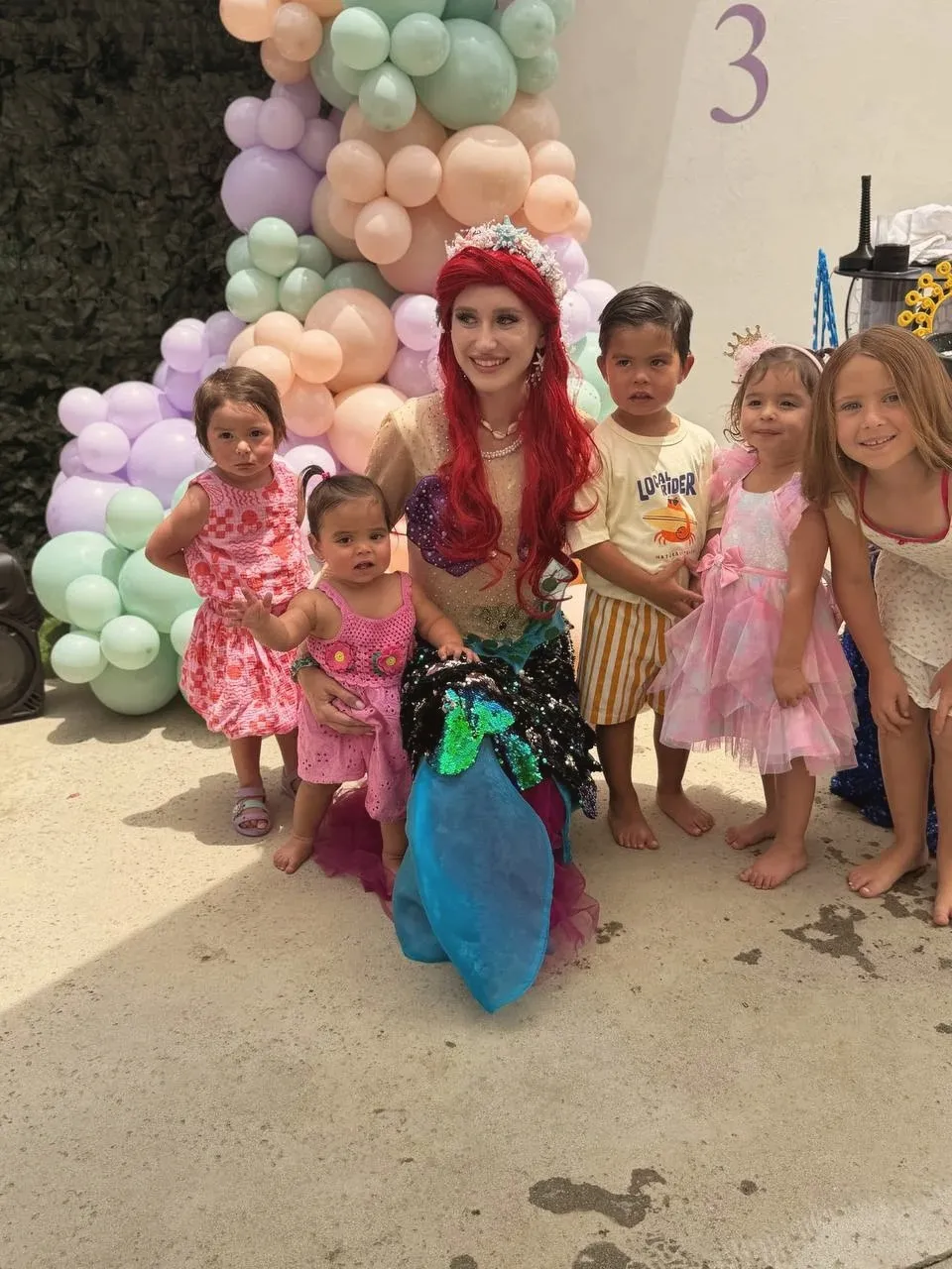 Five small children crowded around the mermaid as she kneels on the patio, her sequinned tail spread out in front of the pastel balloon garland