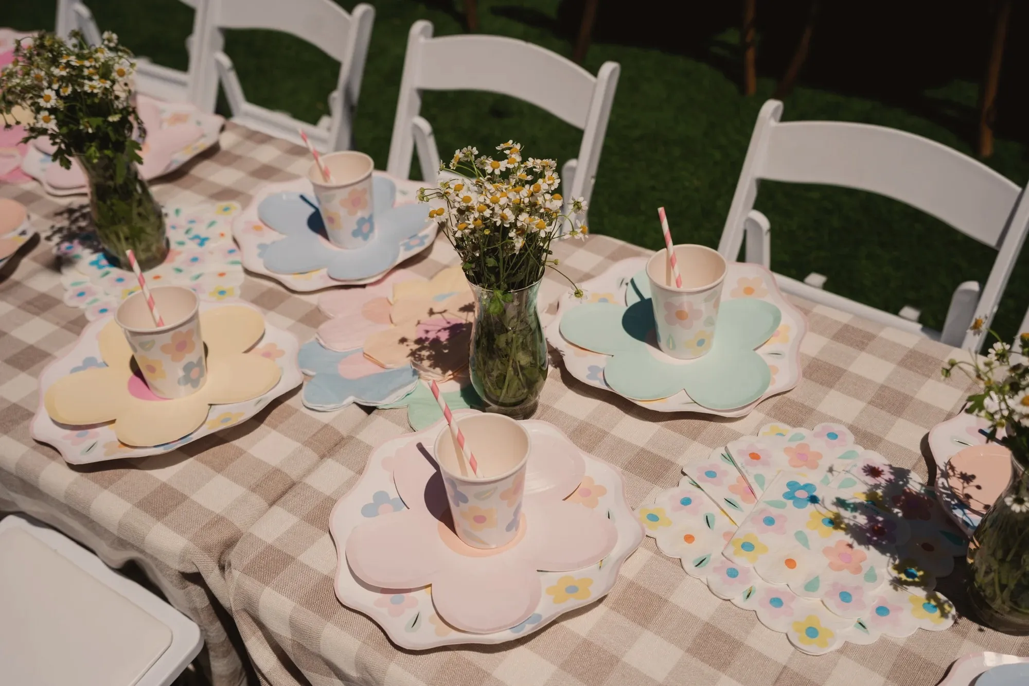 A children's place setting on beige gingham with flower-shaped pastel plates, matching floral cups with striped paper straws and a jar of chamomile