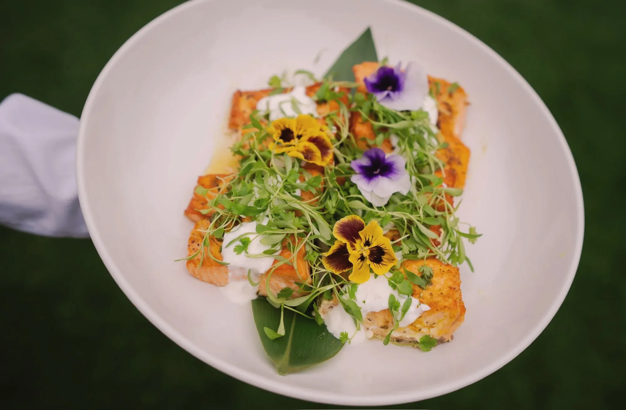 A white bowl of family-style salmon topped with microgreens and yellow and purple edible pansies