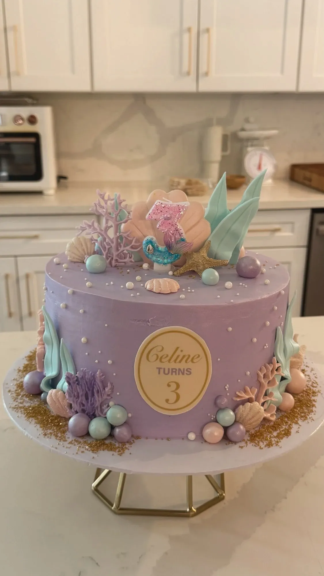 A lilac buttercream cake decorated with fondant coral, shells, pearls and seaweed, topped with a shell and a glittered number three candle, on a gold cake stand