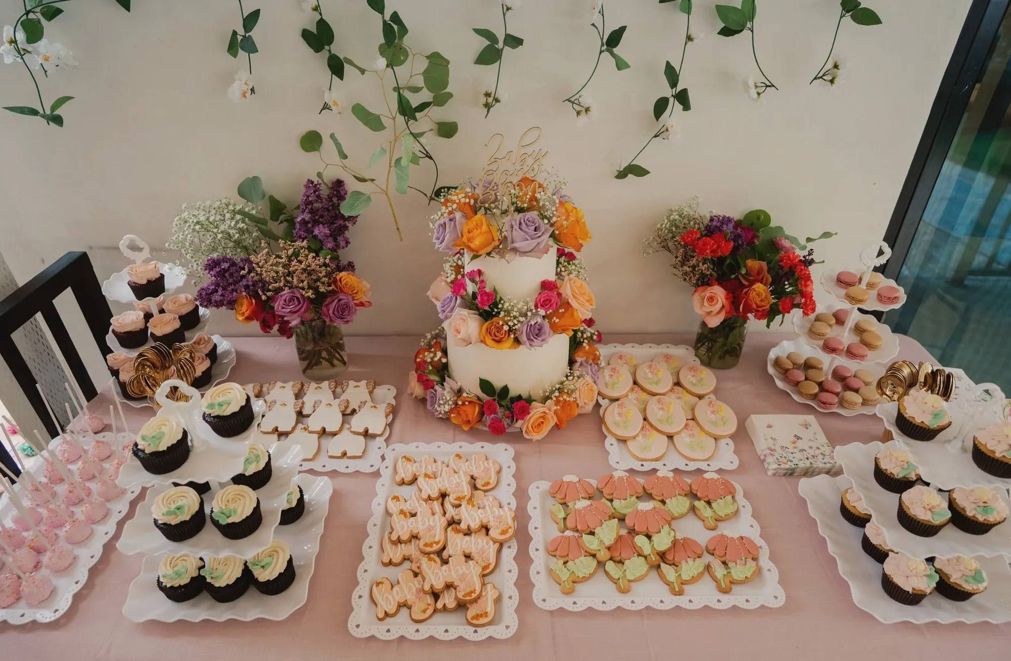 The dessert table in dusty pink, with the flower-wrapped cake in the middle, iced wildflower cookies, macarons and cupcakes on white platters