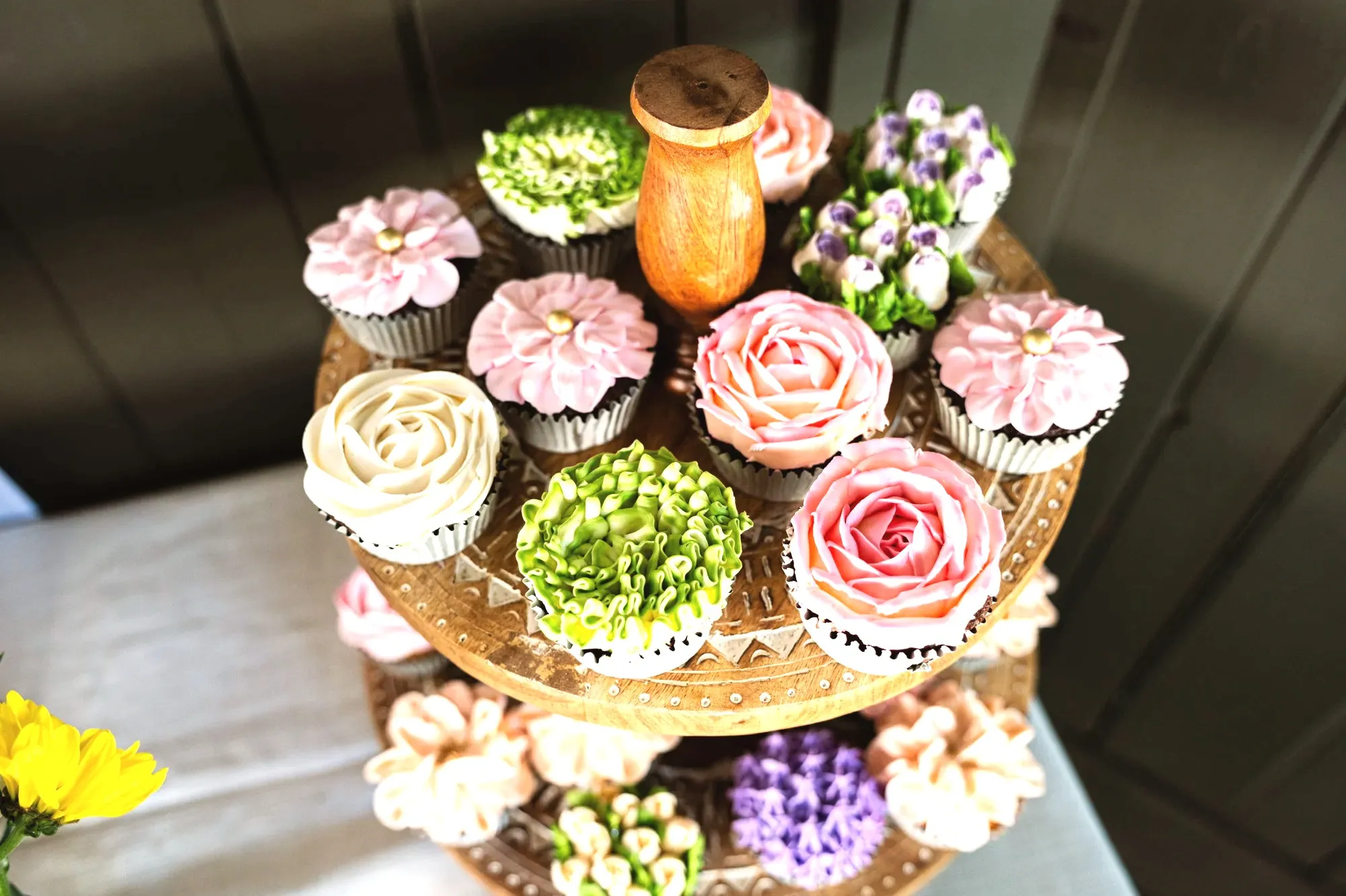 A wooden tiered stand of cupcakes piped as roses, ruffled greenery and lavender sprigs in pink, cream and green buttercream