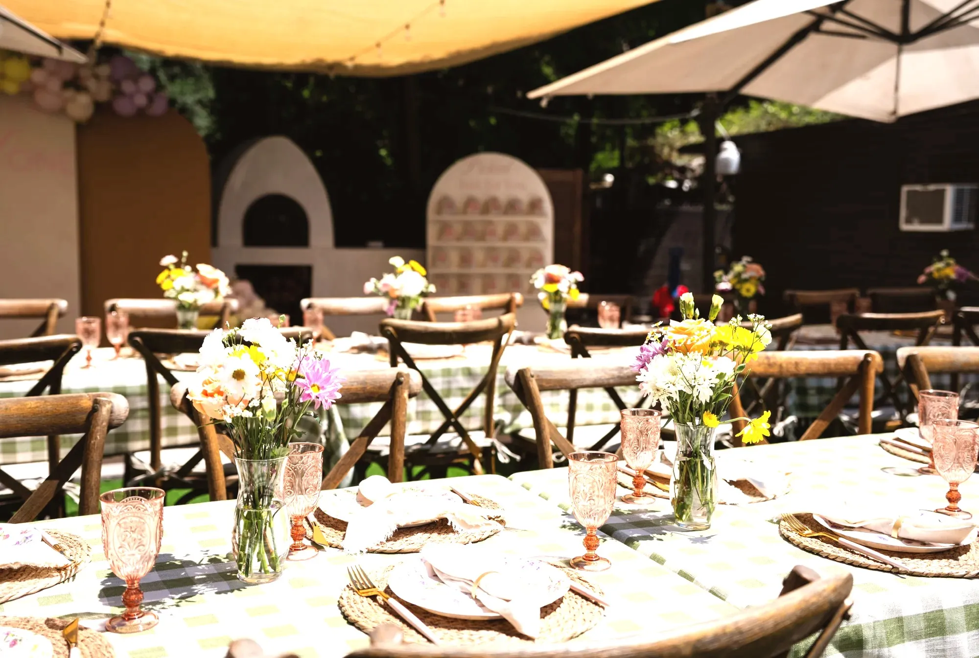 The long gingham tables from the end, with jars of yellow and pink wildflowers, pink pressed-glass goblets and woven placemats under a cream shade sail