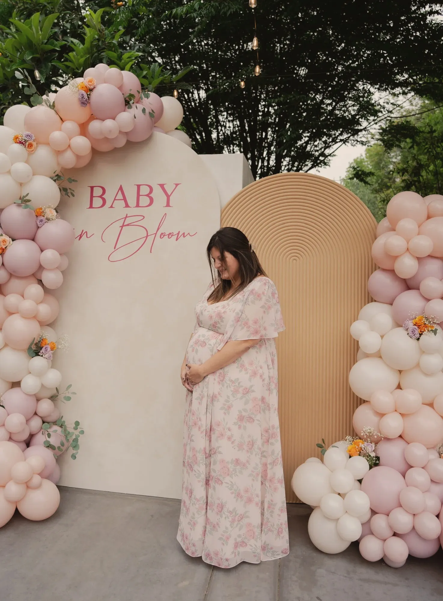 The mum-to-be in a pink floral dress, hands on her bump, in front of the cream Baby in Bloom arch and its pink and lilac balloon garland