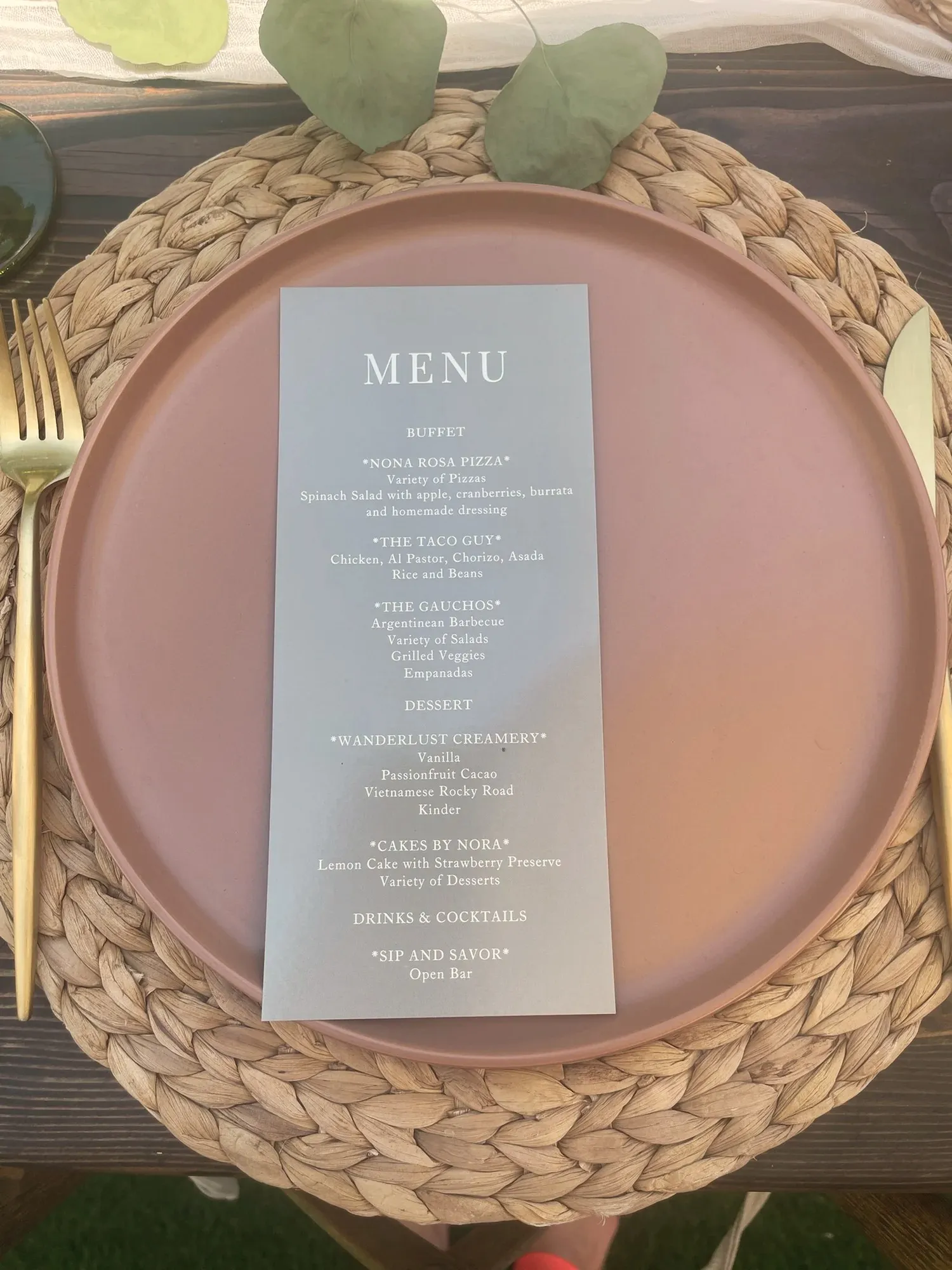 A printed menu card on a terracotta plate and woven placemat, listing the party's three caterers and its dessert and drink stations