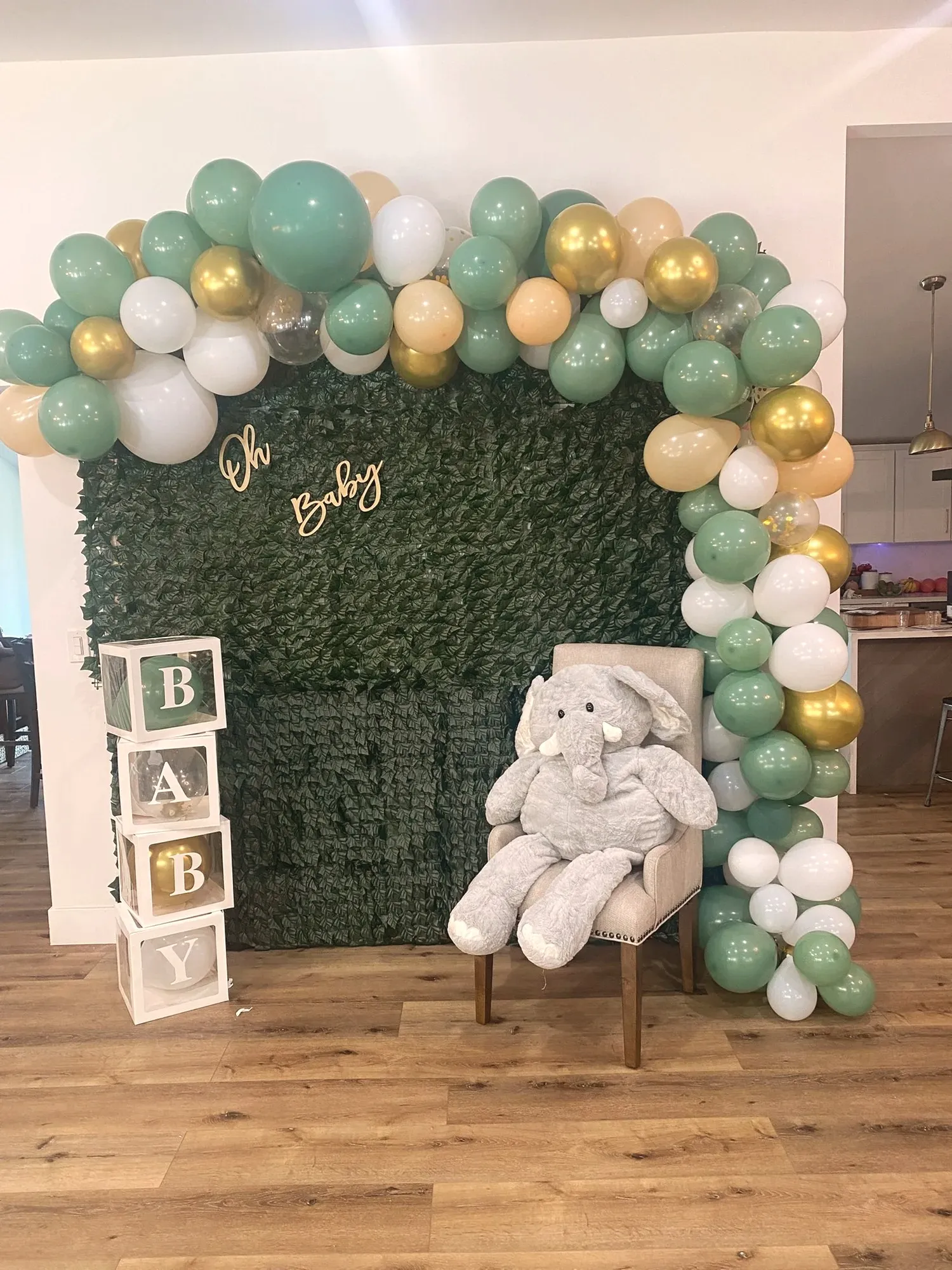 A greenery photo wall with a sage, gold, and white balloon arch, BABY letter blocks, and a large plush elephant on a chair