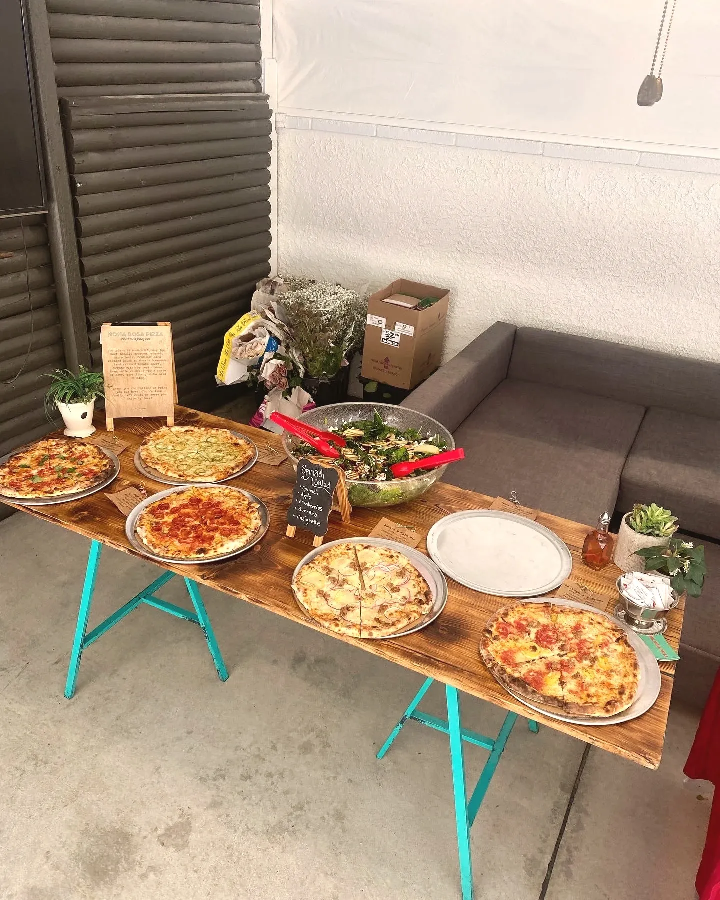 A rustic wood table lined with wood-fired pizzas and a spinach salad, with a handwritten Nona Rosa Pizza sign