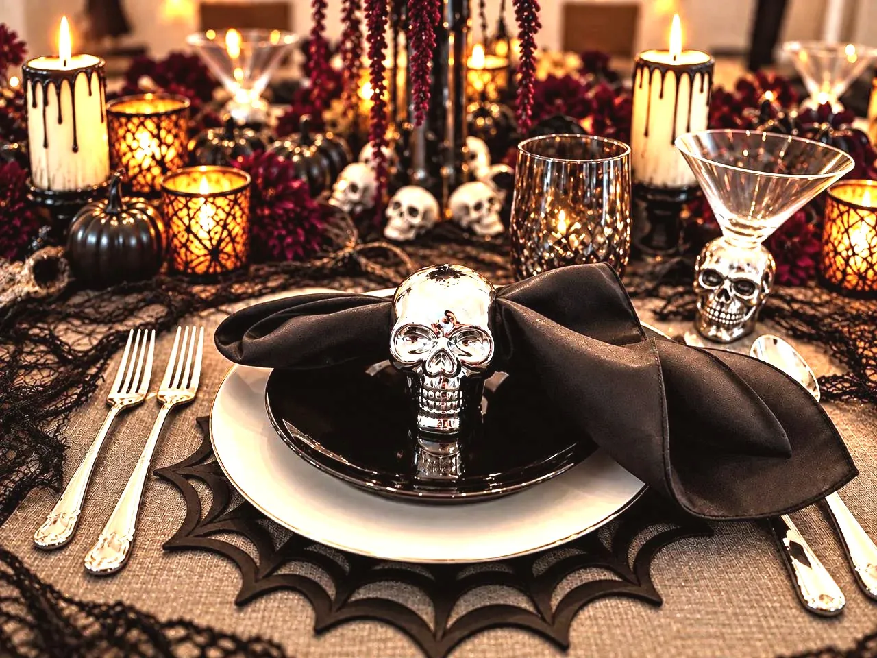 A close place setting with a black napkin through a chrome skull ring on a black plate, a white charger on a black spiderweb placemat, and candles and red flowers behind