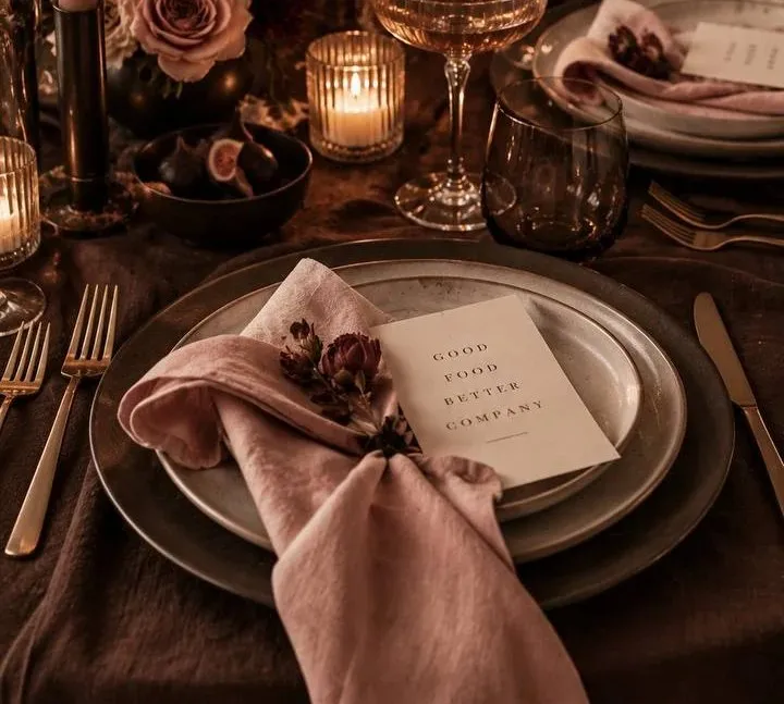 A dining table covered in crumpled deep brown linen, set with a charcoal stoneware charger under a cream speckled dinner plate. A dusty pink linen napkin is knotted loosely across the plate with dried burgundy blooms tucked into the knot, and an ivory card printed "GOOD FOOD BETTER COMPANY" in brown serif capitals rests on top. Brass forks lie to the left and a brass knife to the right, with a ribbed coupe of pale cocktail, a smoked amber tumbler and ribbed glass votives burning just beyond. A pale taper stands in a dark brass holder at the top left beside a small dark bowl of halved figs and the lower blooms of a mauve and blush rose arrangement, and a second setting with a stacked plate, blush napkin and small white place card sits at the top right in the candlelight.